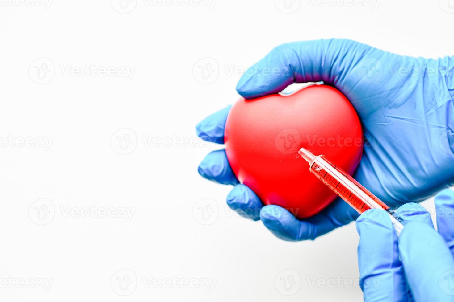 Die Hand des Arztes trug einen blauen Handschuh und hielt ein Herz und eine rote Spritze. Konzept der Koronarangiographie cag. Nahaufnahme, Kopierbereich links für Design oder Text, unscharfer weißer Hintergrund foto