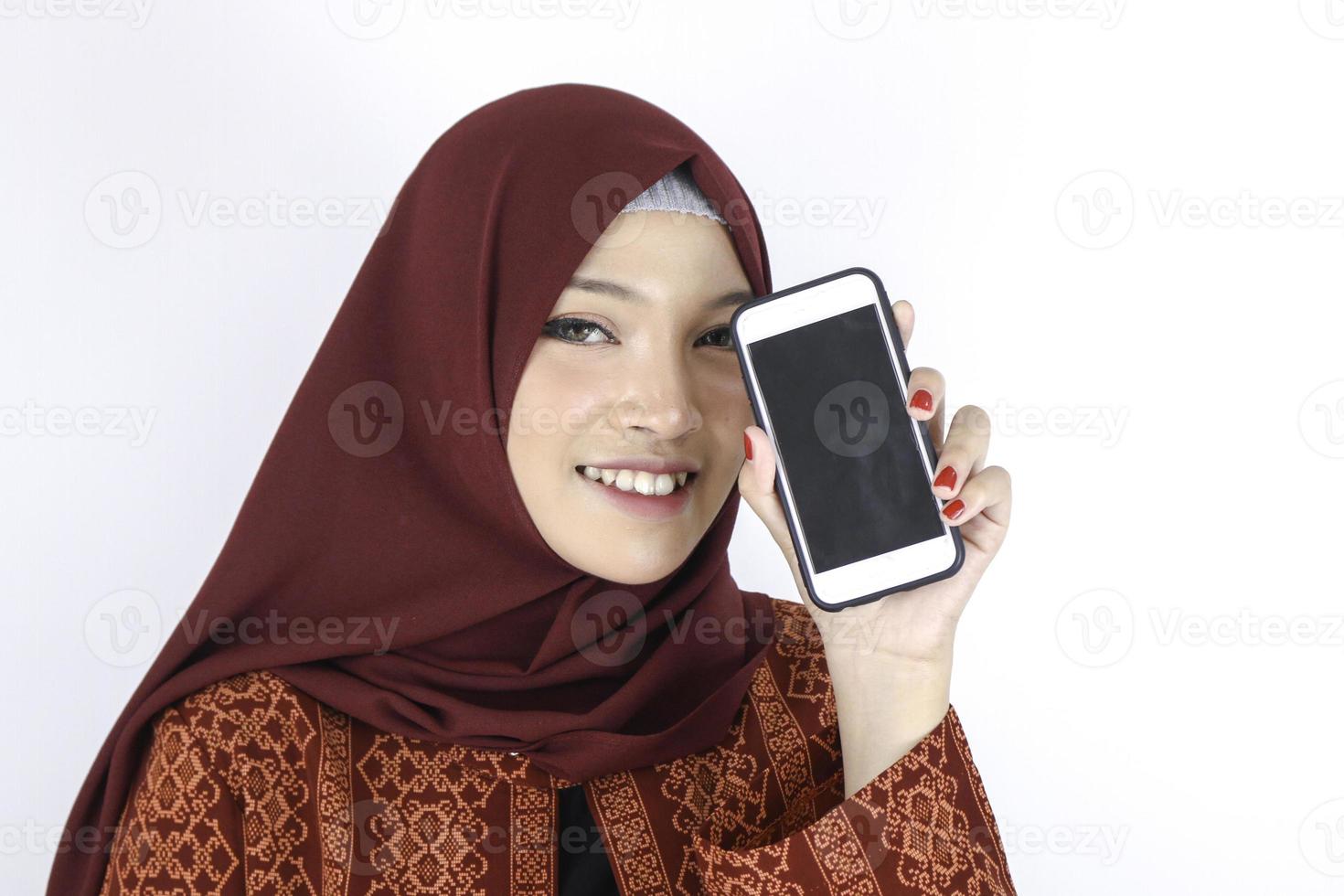 Junge asiatische Islamfrau lächelt und zeigt Smartphone auf weißem Hintergrund. foto