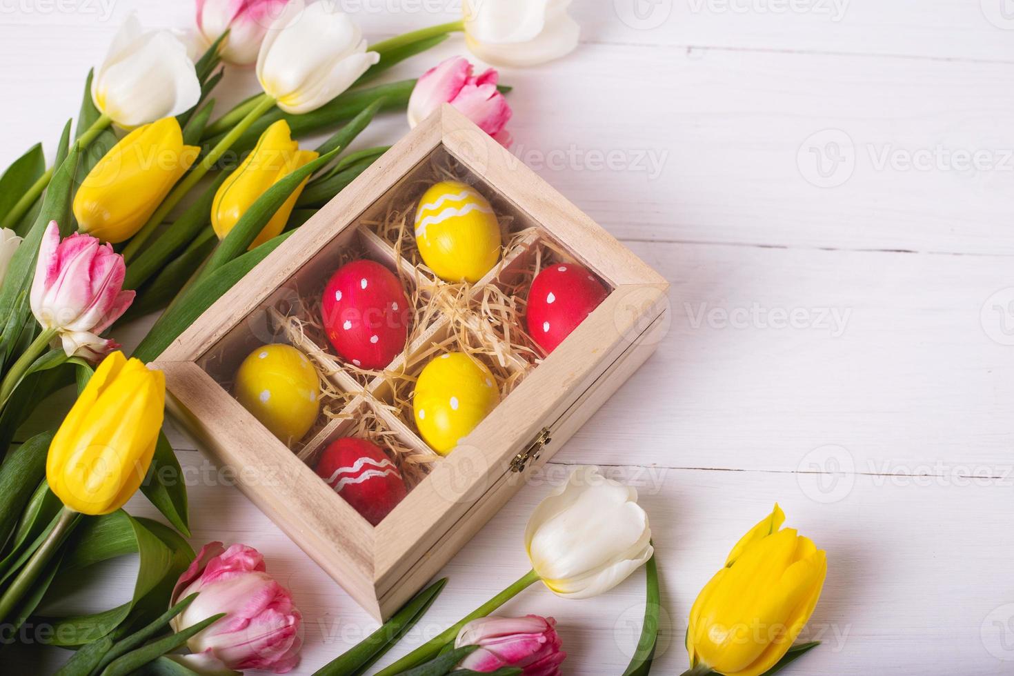 Draufsicht auf bunte Ostereier auf einem Strohbett in einer langen Holzkiste auf einem weißen Holztisch und Tulpen, Platz für Text foto