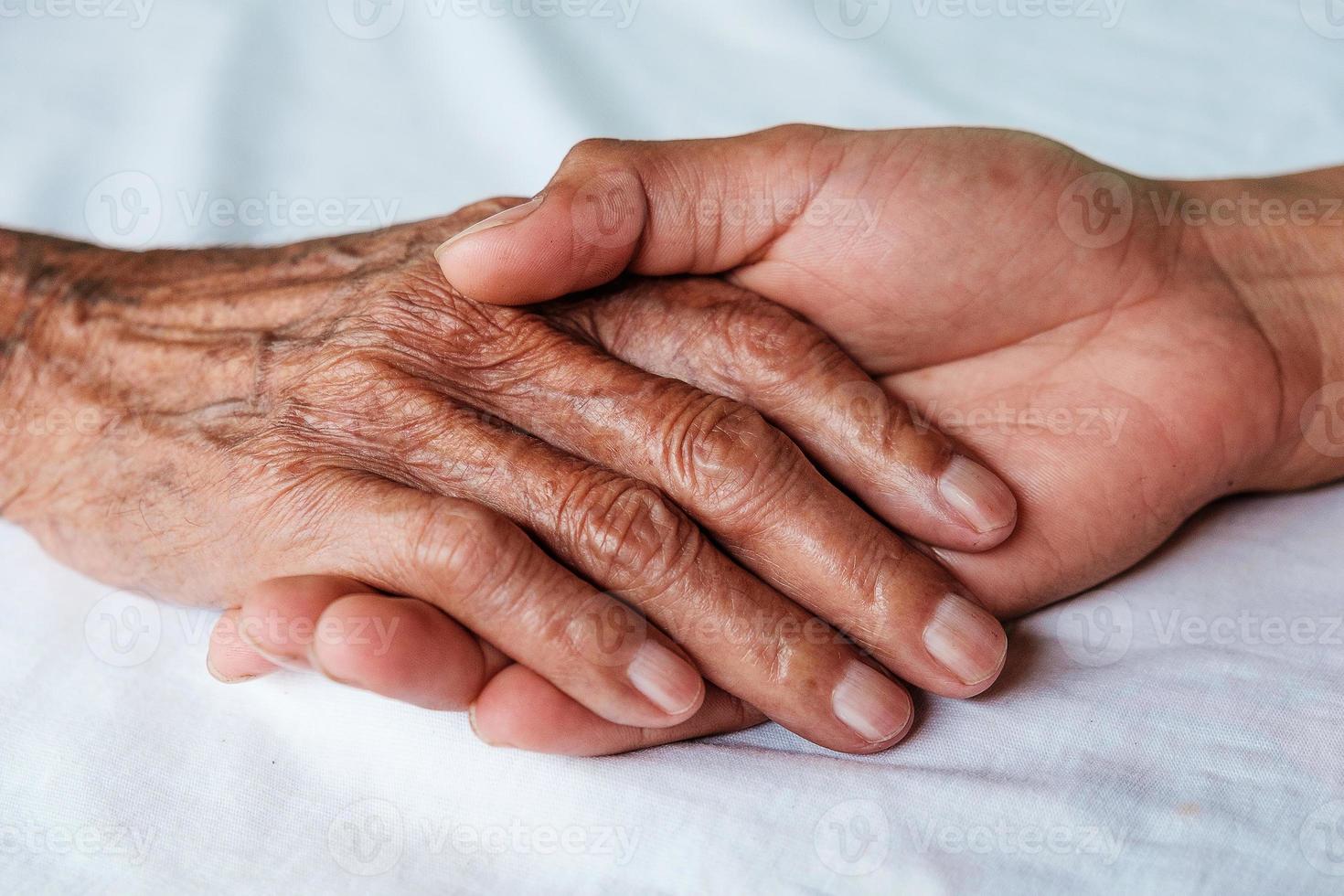 Hände des alten Mannes und eine Männerhand auf dem weißen Bett foto