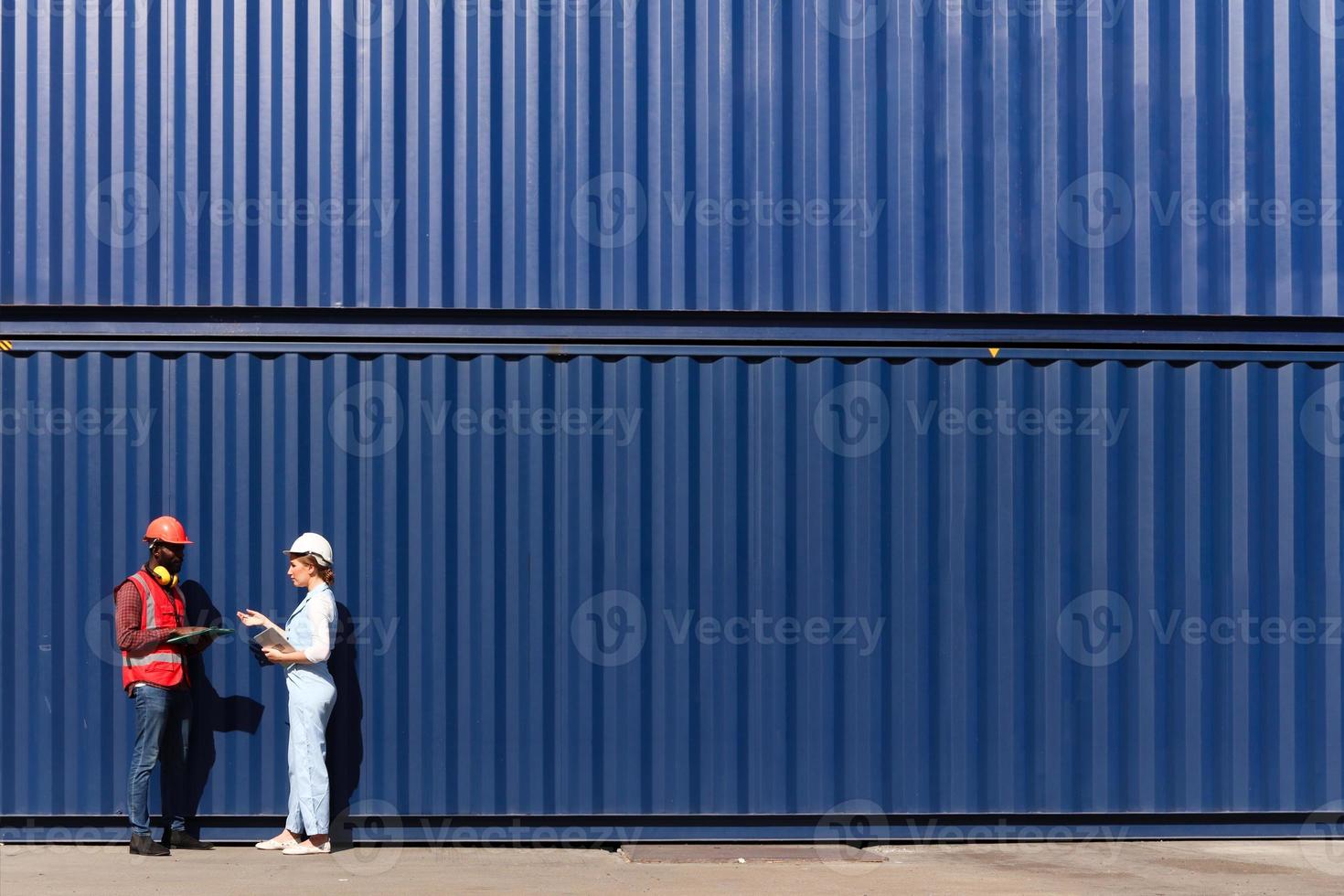 zwei arbeiter tragen sicherheitsweste und helmdiskus am arbeitsplatz des logistikschiffsfrachtcontainerhofs. afrikanisch-amerikanischer ingenieurmann spricht mit schöner frau am blauen containerhintergrund-kopienraum. foto