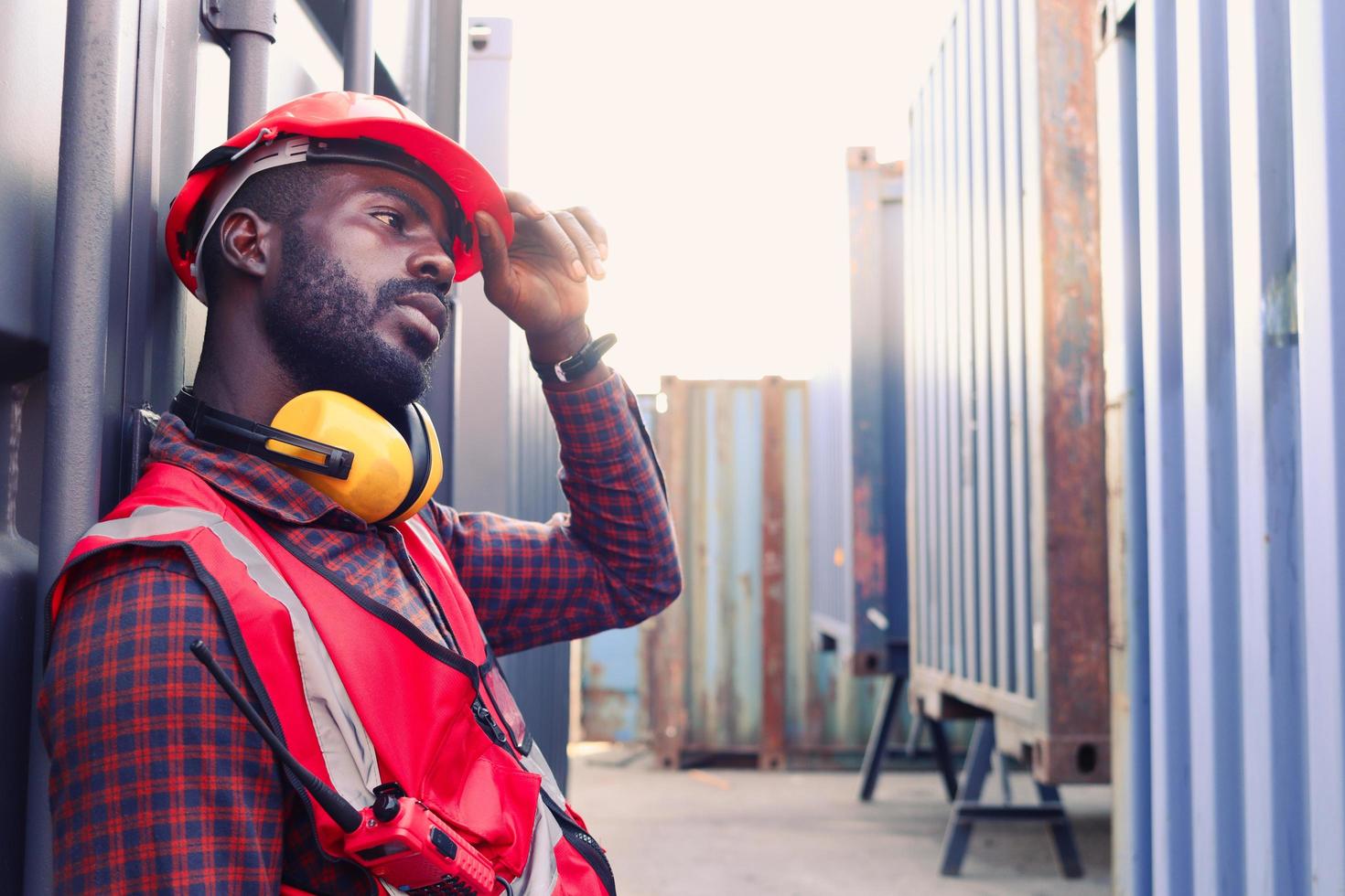 porträt eines afroamerikanischen jungen ingenieurarbeiters trägt eine rote sicherheitsweste und einen helm, fühlt sich müde von der harten arbeit und ruht neben dem versandcontainer auf dem logistischen frachthof. foto