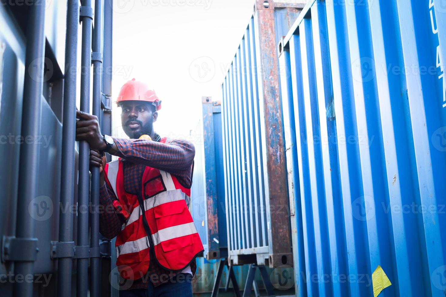 porträt eines afroamerikanischen jungen ingenieurarbeiters, der eine sicherheitsweste und einen helm in neonroter farbe trägt und versucht, eine schiffscontainertür auf dem logistischen frachtcontainerhof zu öffnen. foto