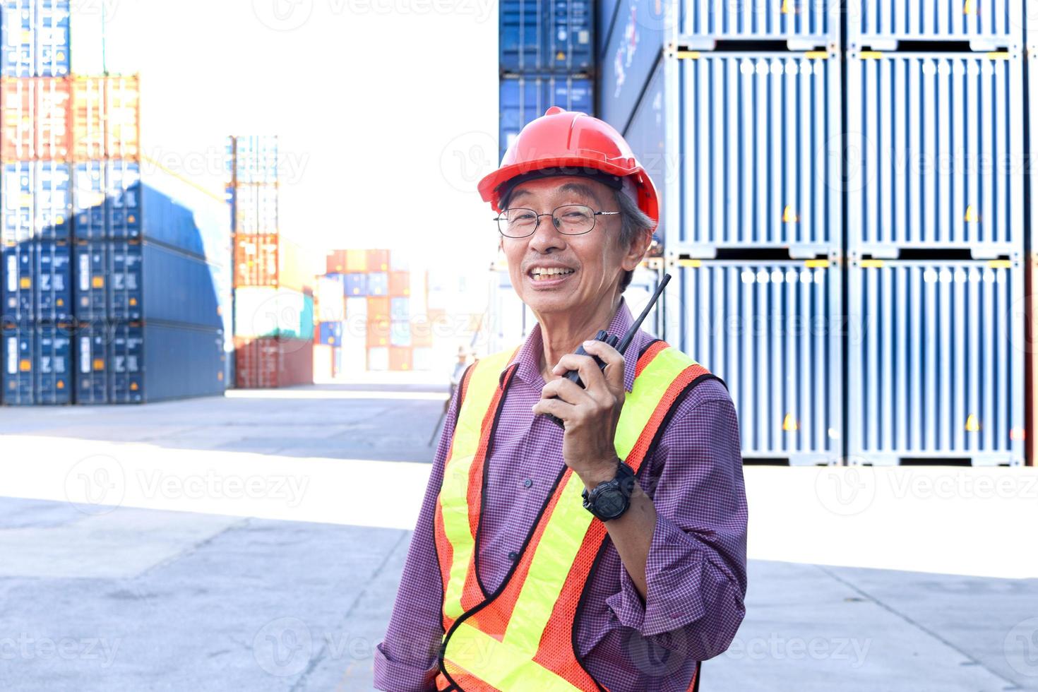 ein hochrangiger älterer asiatischer arbeiter, der sicherheitsweste und helm trägt und funk-walkies-talkie auf dem hof der logistischen frachtcontainer hält. ältere menschen am arbeitsplatzkonzept. foto