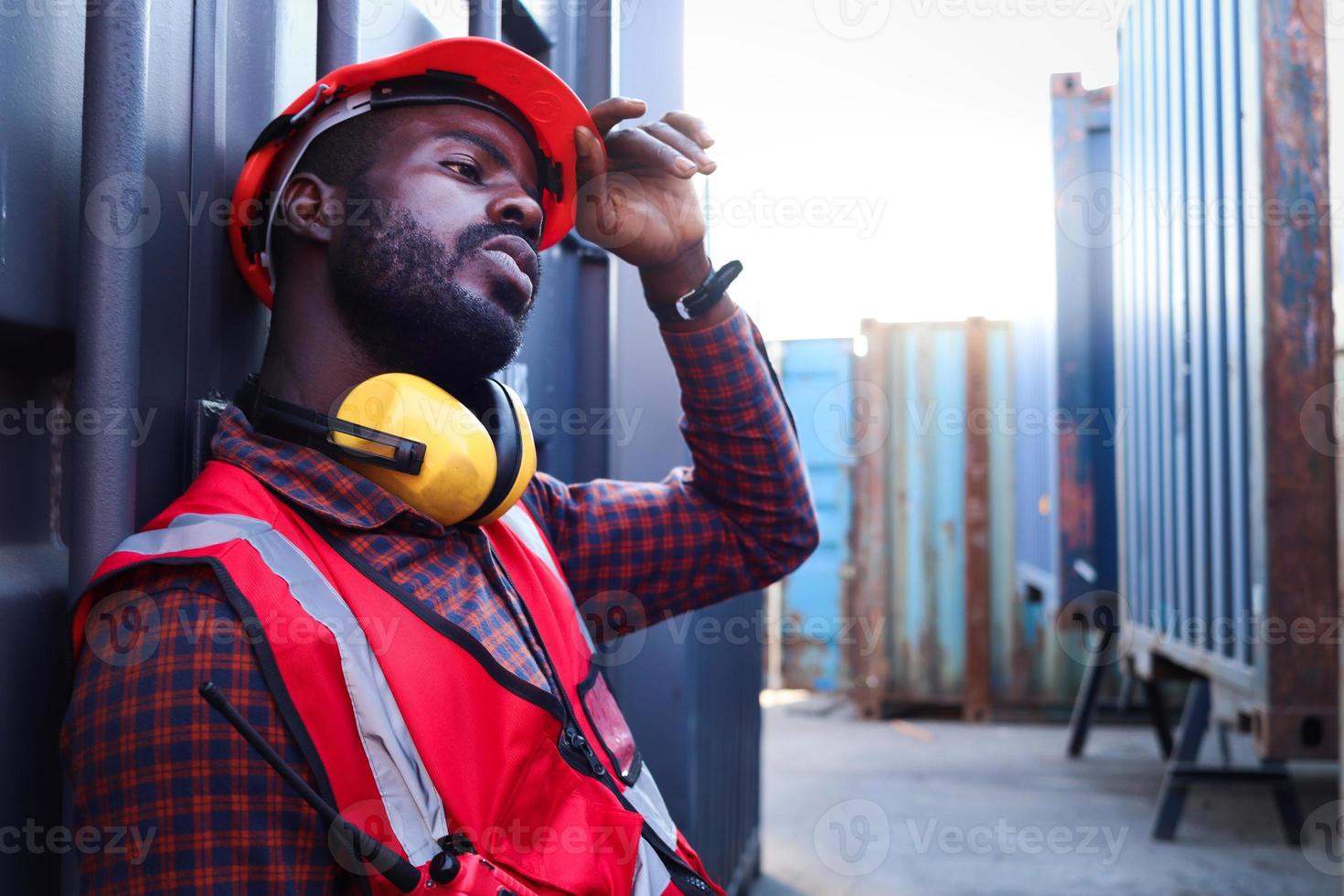 porträt eines afroamerikanischen jungen ingenieurarbeiters trägt eine rote sicherheitsweste und einen helm, fühlt sich müde von der harten arbeit und ruht neben dem versandcontainer auf dem logistischen frachthof. foto