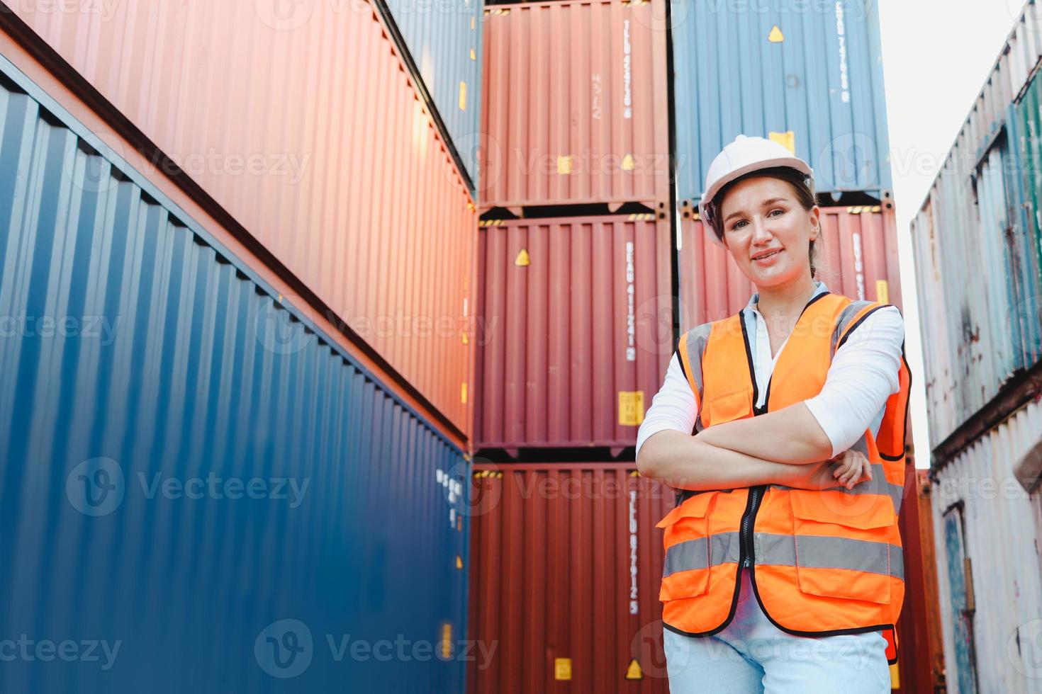 Porträt einer glücklich lächelnden jungen, schönen Ingenieurin mit blonden Haaren, die Sicherheitsweste und Helm trägt und mit verschränktem Arm auf dem Hof der Logistikschifffahrt steht. foto