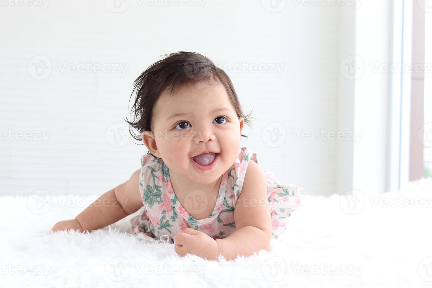 Porträt eines sechs Monate krabbelnden Babys auf flauschigem weißem Teppich, glücklich lächelndes, entzückendes, süßes kleines Mädchen, das im Schlafzimmer auf dem Bett liegt, Kindheits- und Babypflegekonzept foto