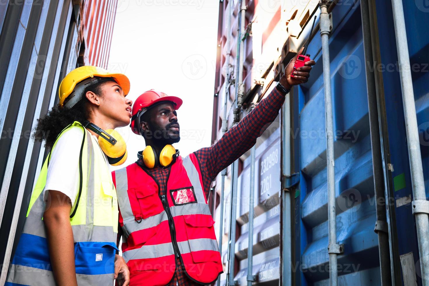 zwei industrielle afroamerikanische ingenieure, mann und frau, die sicherheitsweste und helm tragen, arbeiten zusammen auf dem lager für logistikschiffsfrachtcontainer, arbeitermann, der walkie-talkie hält und wegzeigt. foto