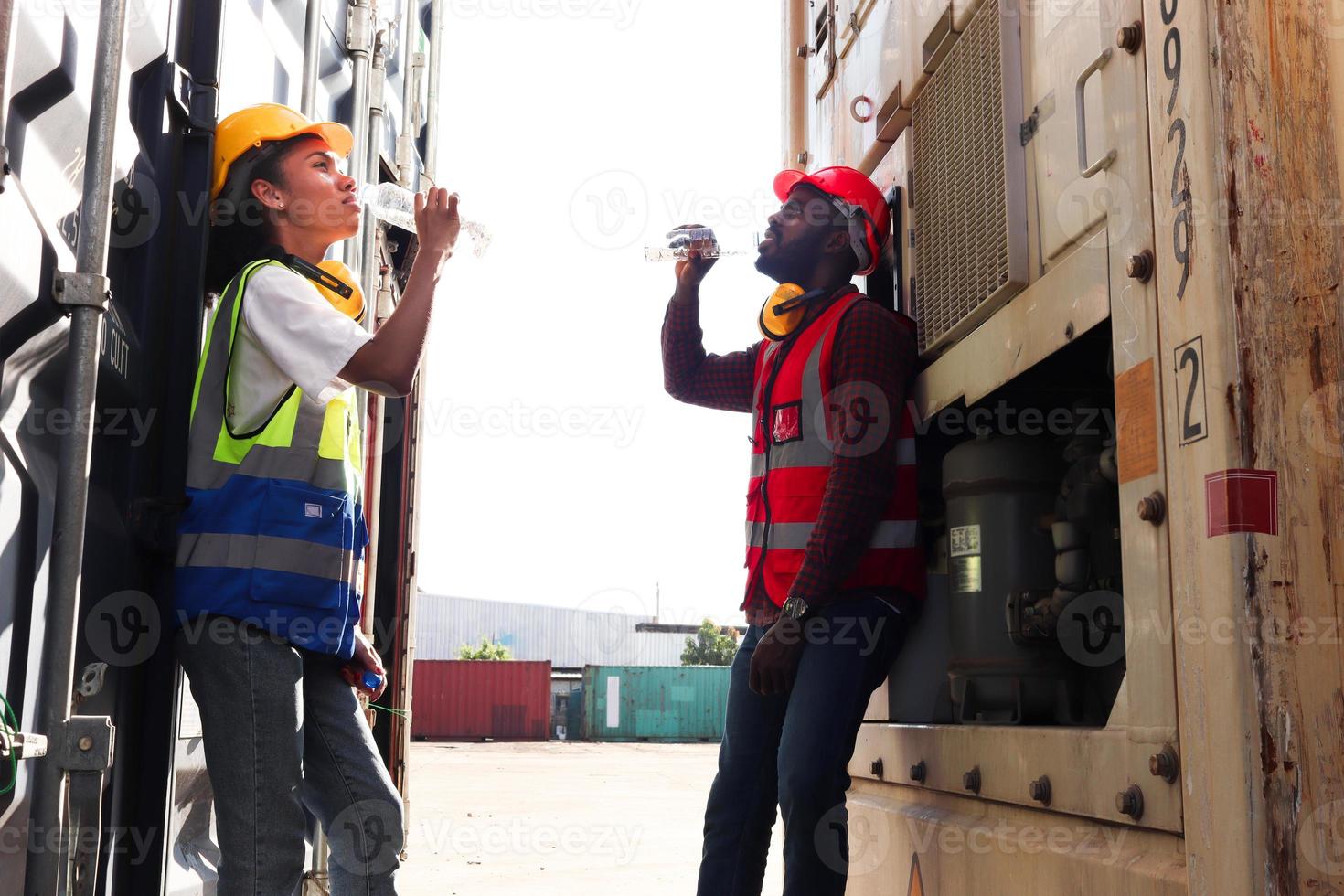 zwei industrielle afroamerikanische ingenieure, mann und frau, die sicherheitsweste und helm tragen, trinken wasser aus der flasche während einer pause nach der harten arbeit auf dem hof der logistischen frachtcontainer. foto