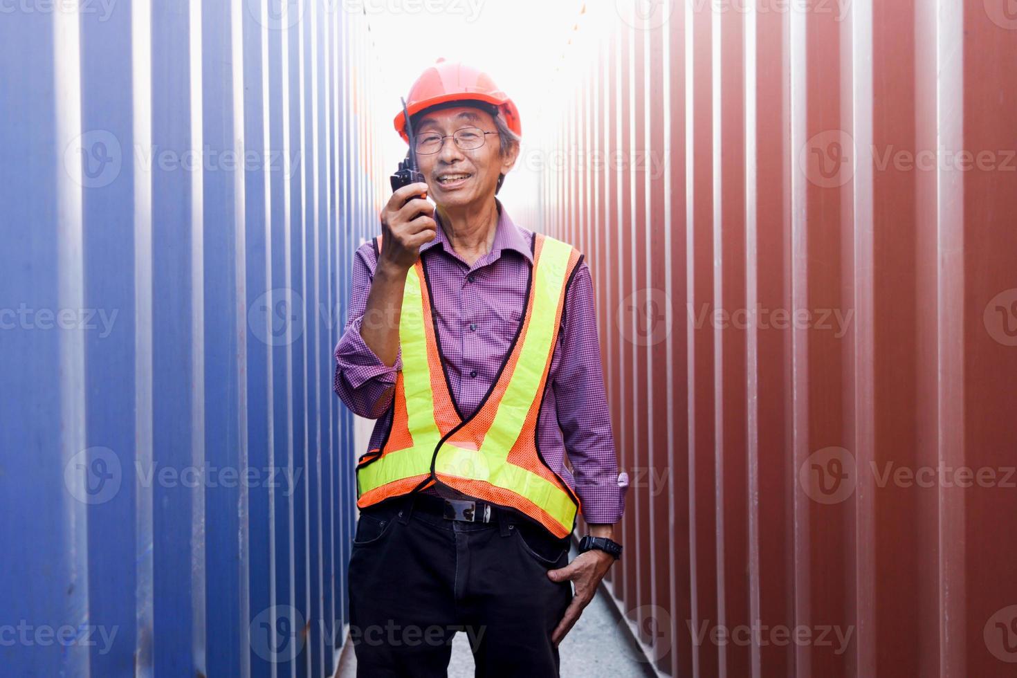 porträt eines älteren asiatischen arbeitsingenieurs mit sicherheitsweste und helm, der ein radio-walkie-talkie hält und zwischen roten und blauen containern auf dem hof der logistischen frachtcontainer steht. foto