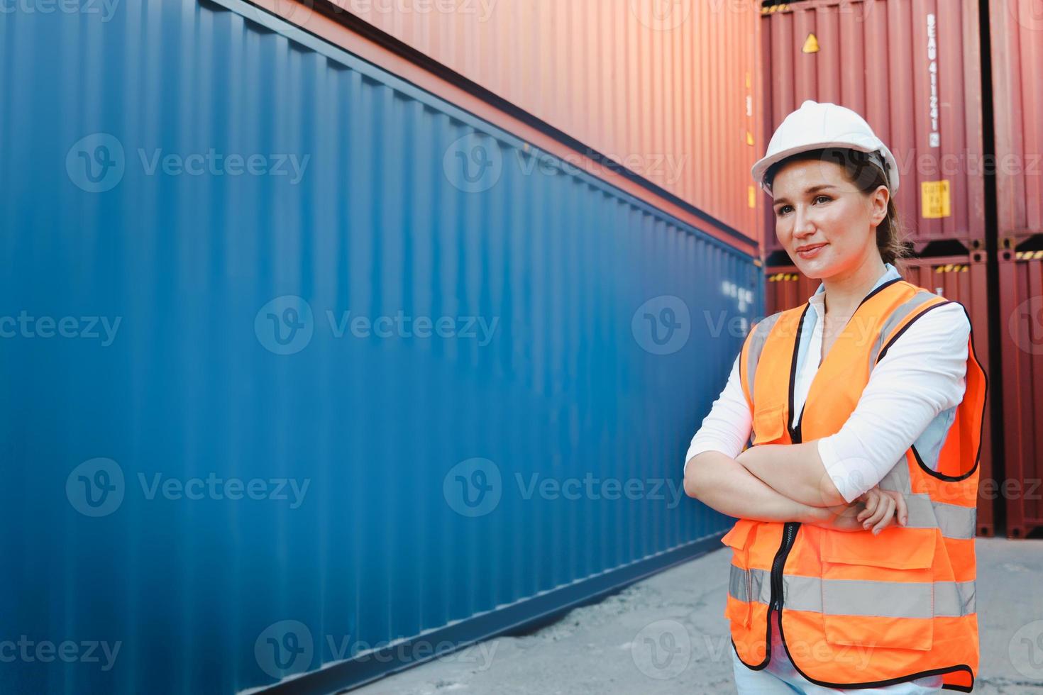 Porträt einer glücklich lächelnden jungen, schönen Ingenieurin mit blonden Haaren, die Sicherheitsweste und Helm trägt und mit verschränktem Arm auf dem Hof der Logistikschifffahrt steht. foto