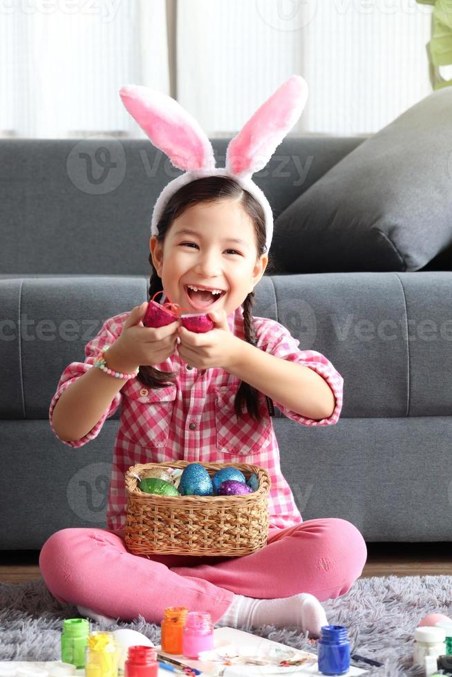 Fröhliches entzückendes rosafarbenes kleines Osterhasenmädchenkind mit Hasenohren-Stirnband, das lächelt und lacht, nachdem es bunte ostereier gefunden hat, Eierjagdspiel, Feier des Frühlingsanfangs. foto