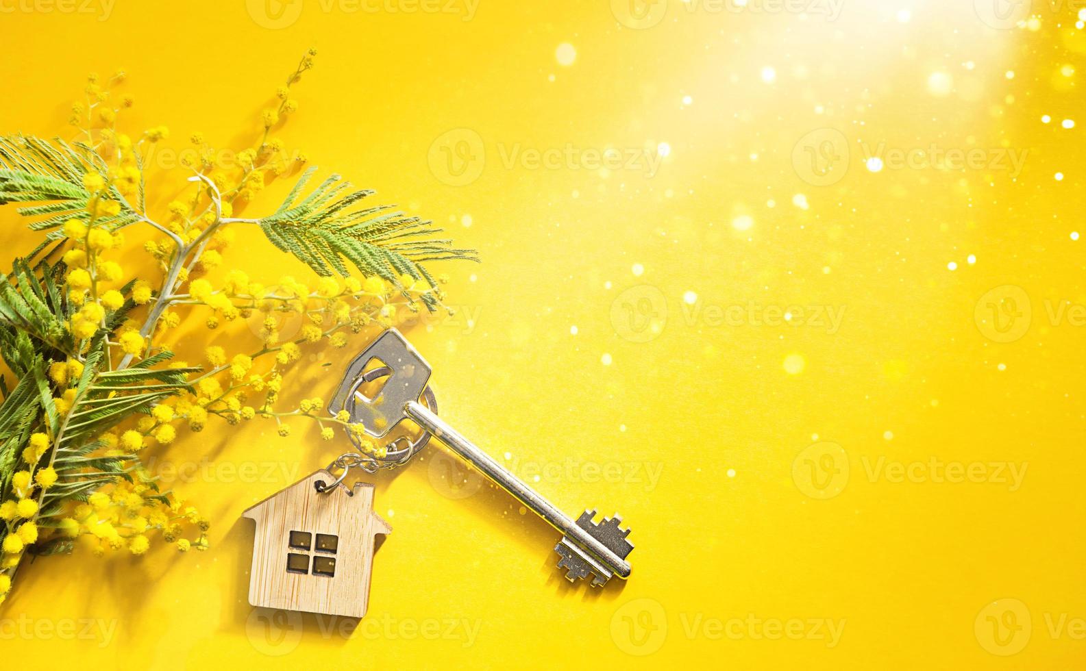 Schlüsselanhänger in Form eines Holzhauses mit Schlüssel auf gelbem Hintergrund und Frühlingsmimose. Bauen, Design, Projekt, Umzug in ein neues Zuhause, Hypothek, Miete und Kauf von Immobilien. Platz kopieren foto