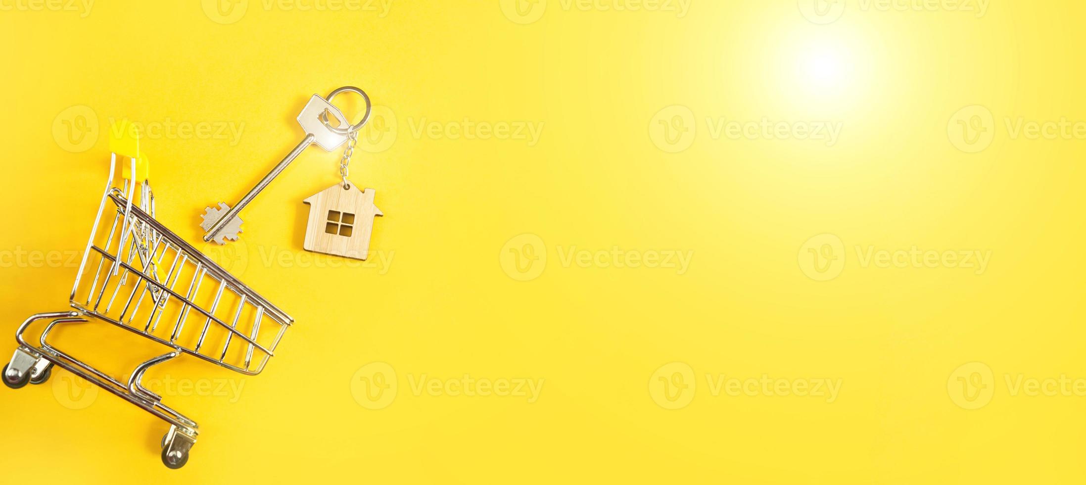 Schlüsselanhänger in Form eines Holzhauses mit Schlüssel auf gelbem Hintergrund und Einkaufswagen. Hypothek, Miete und Kauf von Immobilien, Frühjahrs- und Sommerangebot für den Bau. Platz kopieren foto
