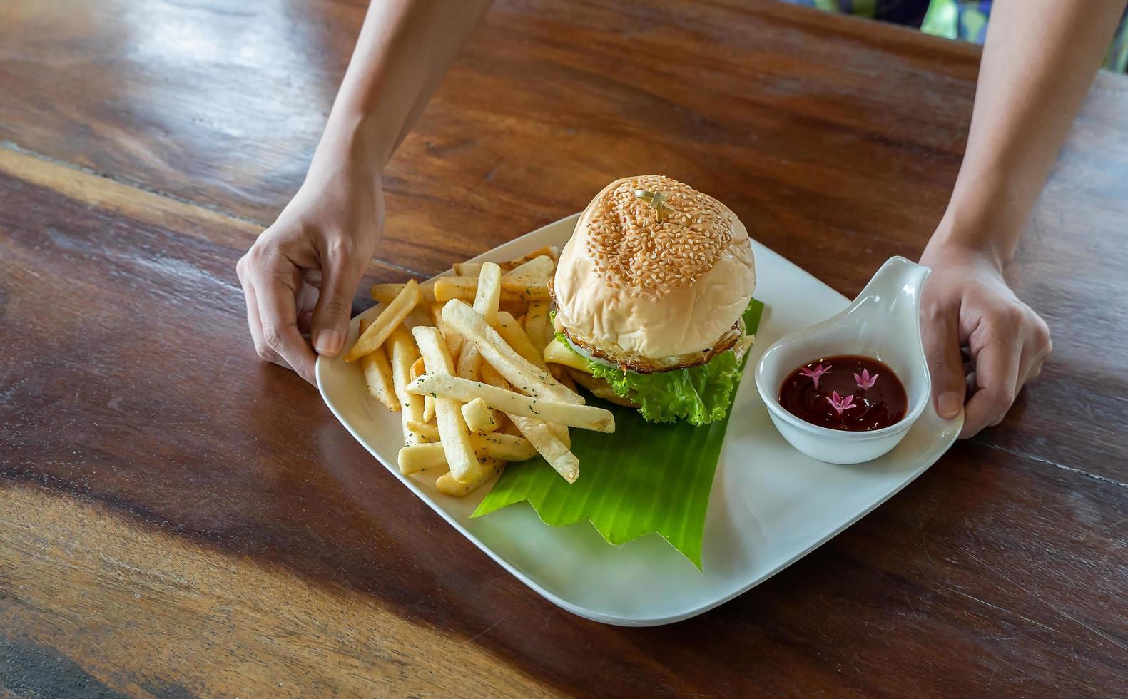 Hände, die einen Hamburger halten, junges Mädchen, das Fast-Food-Burger mit Pommes Frites, Tomatensauce auf einem weißen Teller hält. foto