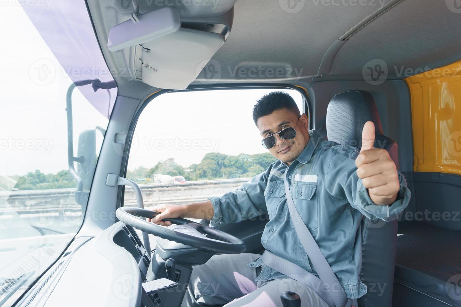 professioneller lkw-fahrer, erwachsener männlicher asiate mit sonnenbrille lächelt und hebt die hand. Der junge Arbeiter vertraut seit langem auf die Sicherheit des Transport- und Liefergeschäfts. foto