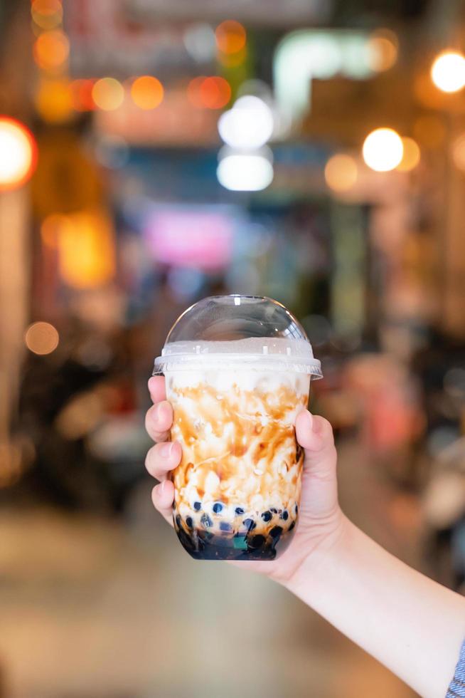 junge frau hält, trinkt braunen zuckergeschmack tapioka perlenblasenmilchtee mit glasstroh auf dem nachtmarkt von taiwan, nahaufnahme, bokeh foto