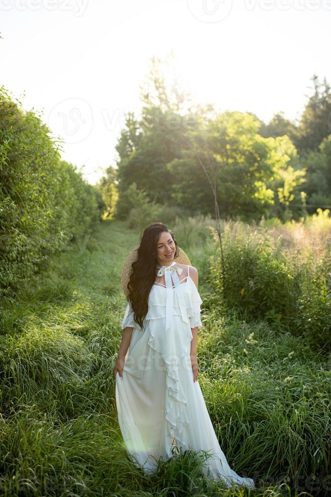 Schwangere Frau mit Hut, die in einem Kleid auf einem Hintergrund von grünen Bäumen posiert. foto