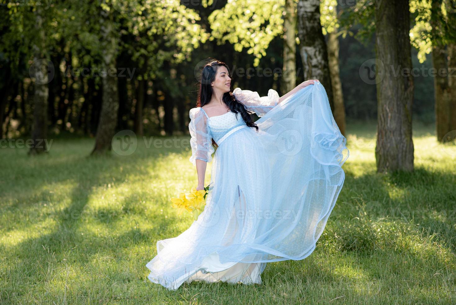 Schwangere Frau mit Hut, die in einem Kleid auf einem Hintergrund von grünen Bäumen posiert. foto