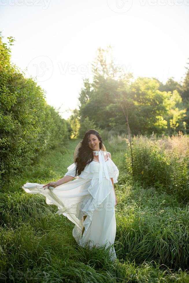 Schwangere Frau mit Hut, die in einem Kleid auf einem Hintergrund von grünen Bäumen posiert. foto