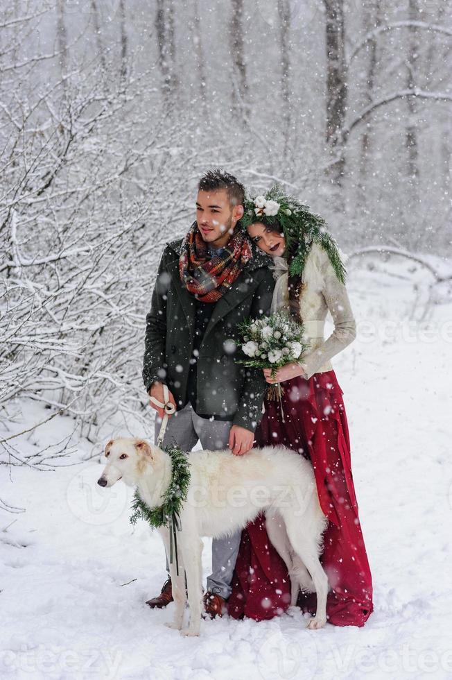 Schöne Braut und Bräutigam mit einem weißen Hund stehen auf dem Hintergrund eines verschneiten Waldes. foto