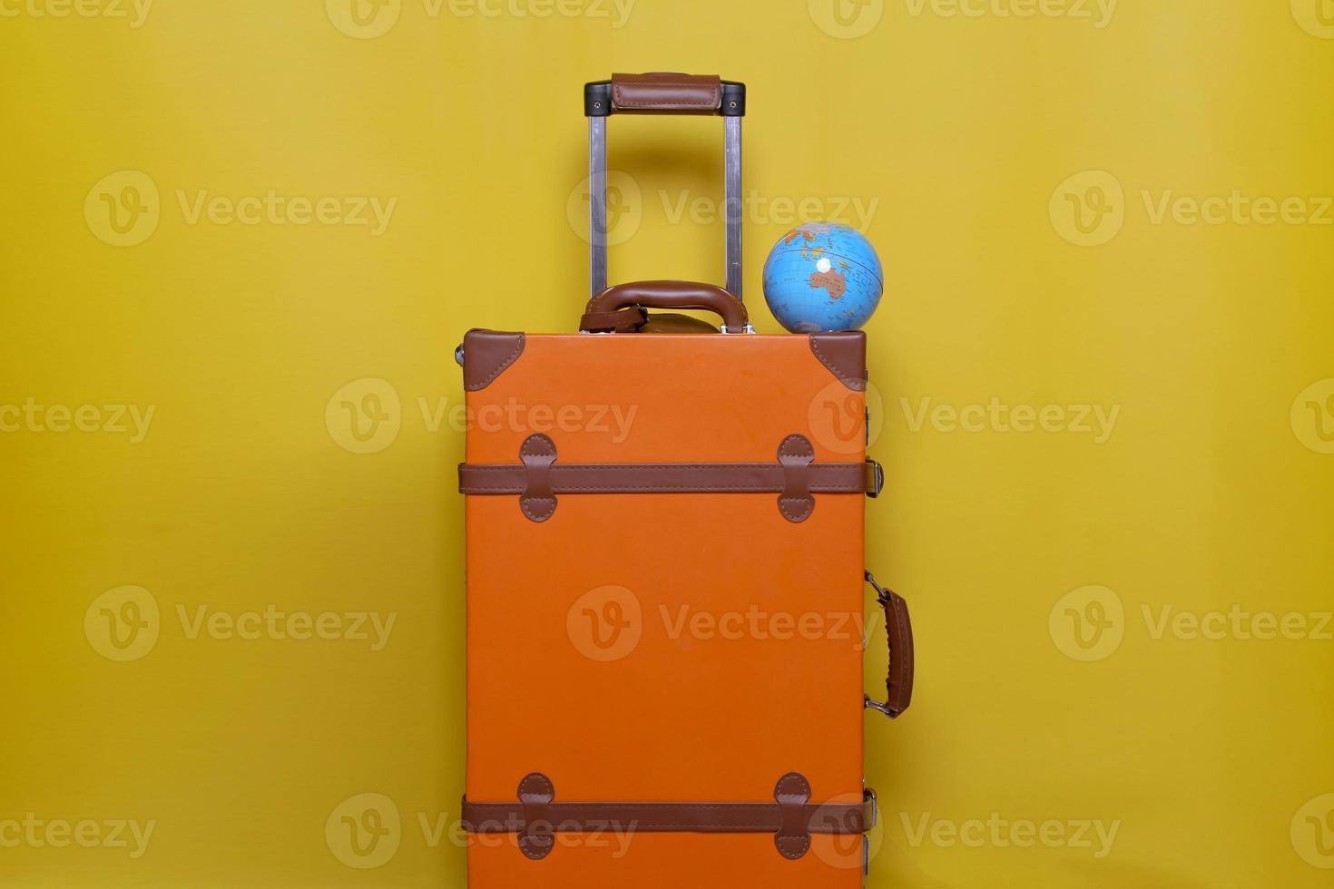 Orangefarbener Koffer mit Mini-Globus isoliert auf gelbem Hintergrund für Reisekonzept mit minimalem Stil foto