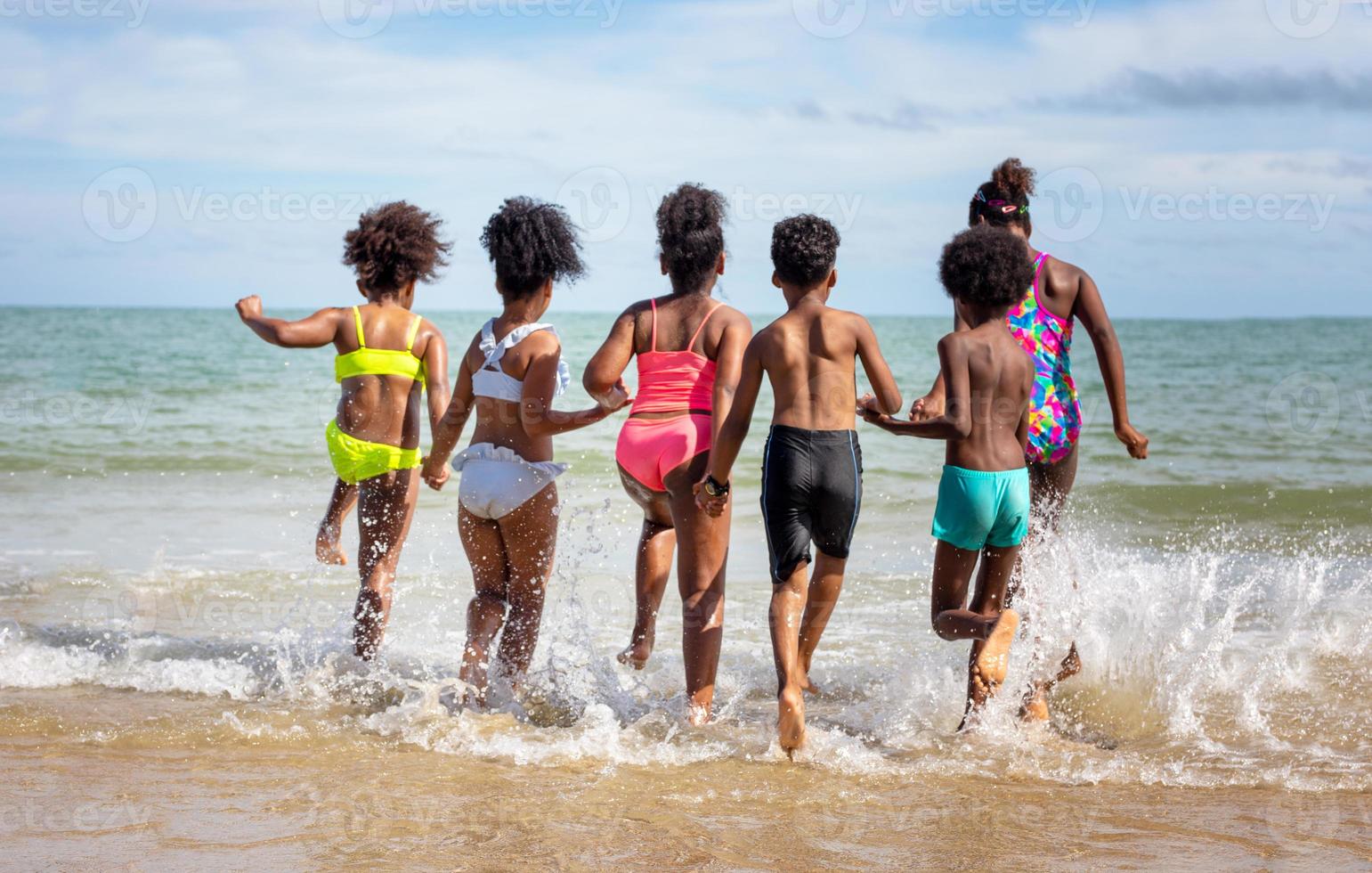 Kinder, die am Strand auf Sand spielen, eine Gruppe von Kindern, die im Sommer am Strand Händchen halten, Rückansicht gegen Meer und blauen Himmel foto