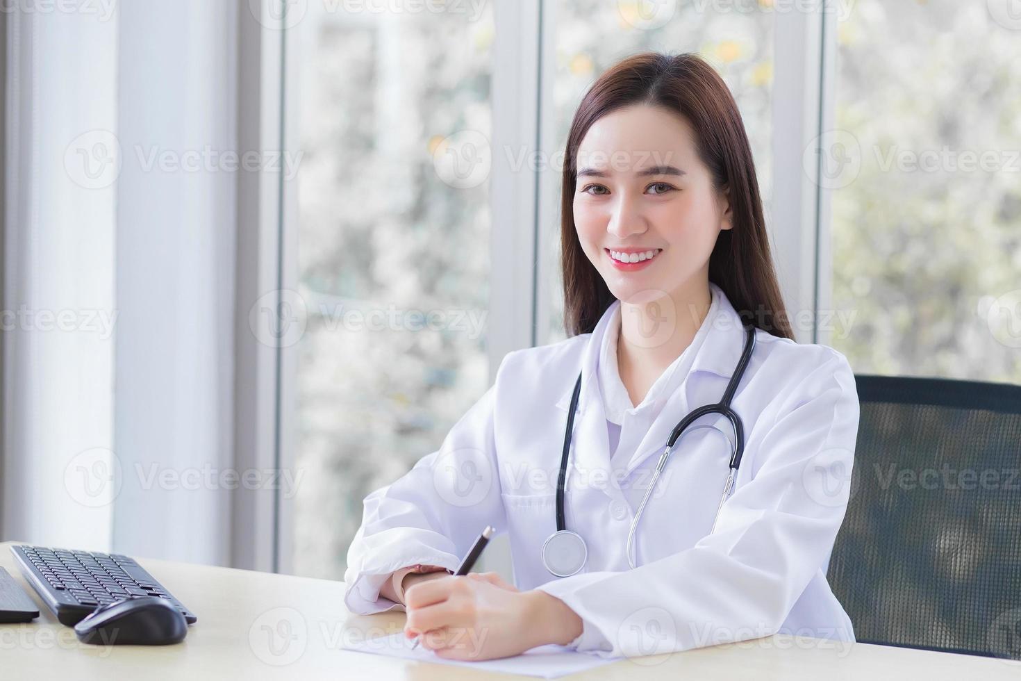 Professionelle asiatische Ärztin trägt Arztkittel und Stethoskop, während sie etwas auf das Papier schreibt und im Büroraum arbeitet, den sie im Krankenhaus in die Kamera schaut. foto