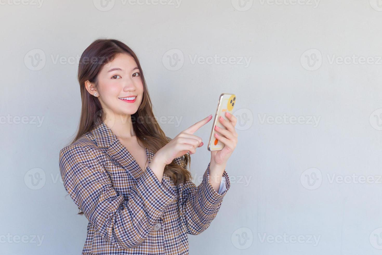 asiatische berufstätige frau mit schwarzen langen haaren, die einen karierten anzug tragen, während sie mit erfolg bei der arbeit genießt und mit einem smartphone auf weißem hintergrund in die kamera schaut. foto