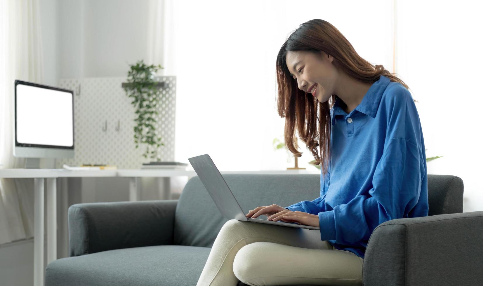 junge freiberufliche asiatische frau, die am laptop arbeitet und soziale medien überprüft, während sie auf dem sofa liegt, wenn sie sich im wohnzimmer zu hause entspannen. foto