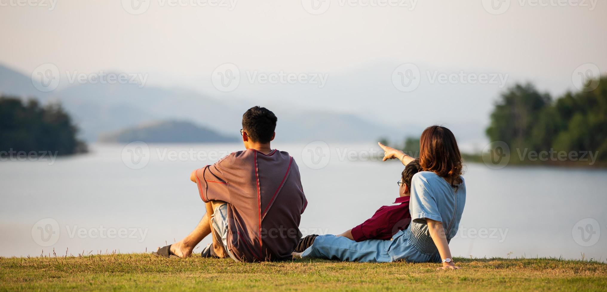 mutter und zwei söhne stehen neben einem großen see und sehen im hintergrund bergblick, mutter zeigt mit dem finger auf den wald. Idee für gemeinsame touristische Familienreisen bis hin zum Outdoor-Trip foto