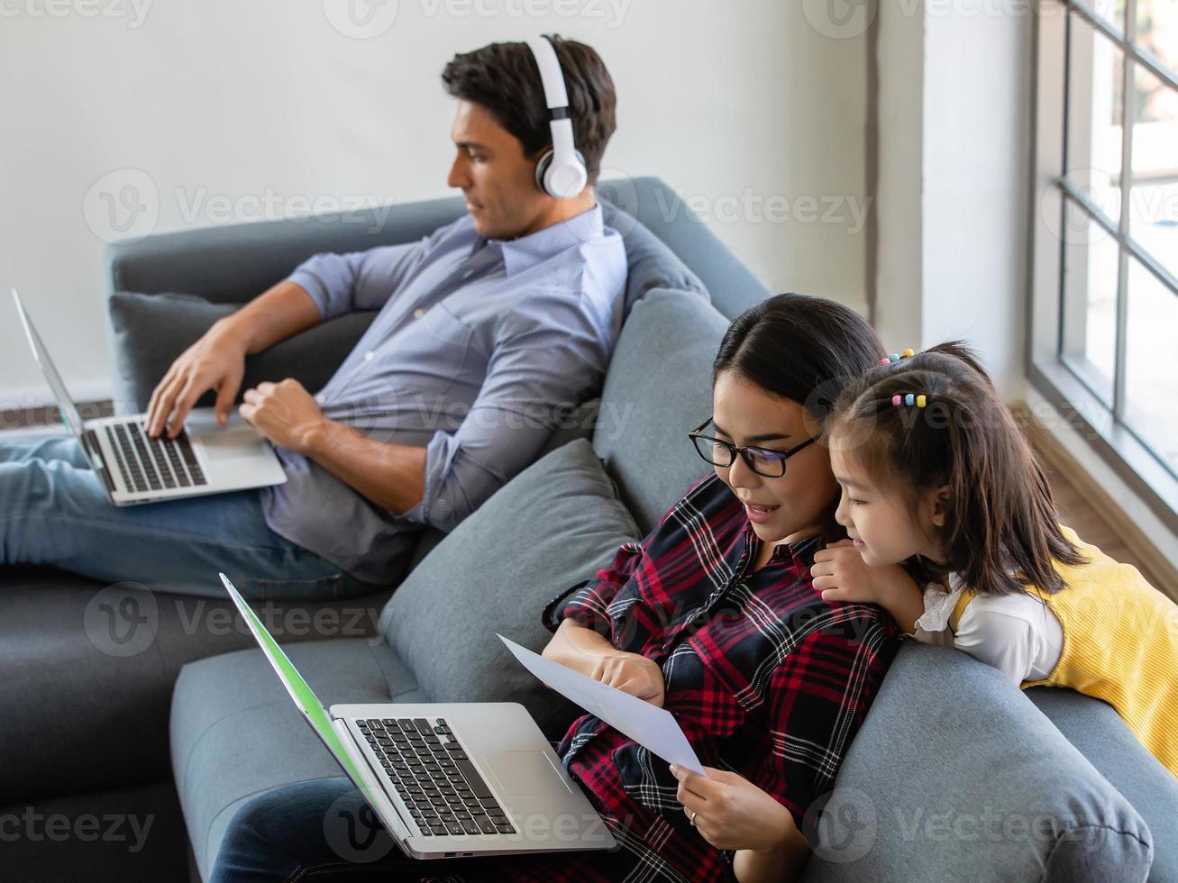 gemischtrassige familie, vater, mutter und kleine tochter bleiben zusammen zu hause. mama und papa arbeiten mit laptop-notizbuch und unterrichten das mädchen. Arbeit von zu Hause aus und neues normales Konzept foto