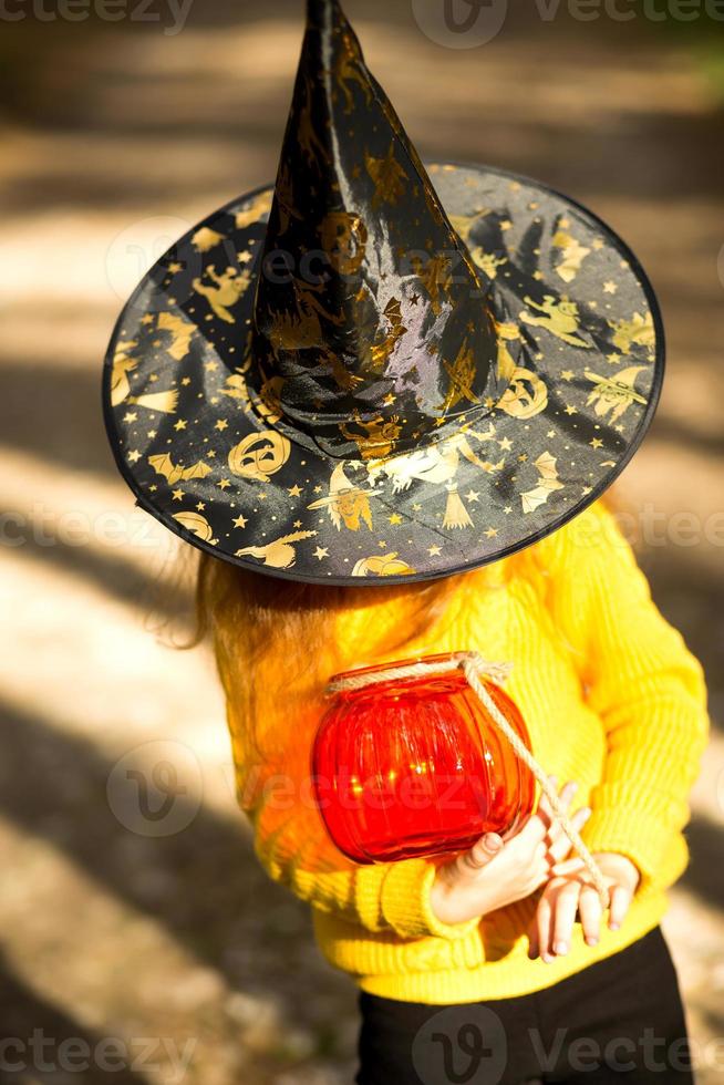 ein Mädchen in einem Hexenhut, in einem gelben Pullover und mit einem Kürbis, einer Laterne und einem Eimer für Süßigkeiten im Herbstwald. Halloween-Urlaub. Porträt, Nahaufnahme foto