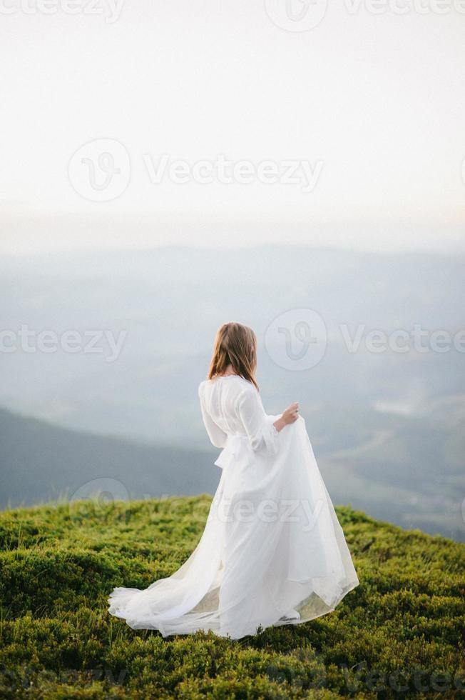 Frau im Hochzeitskleid läuft über das Feld in Richtung der Berge foto