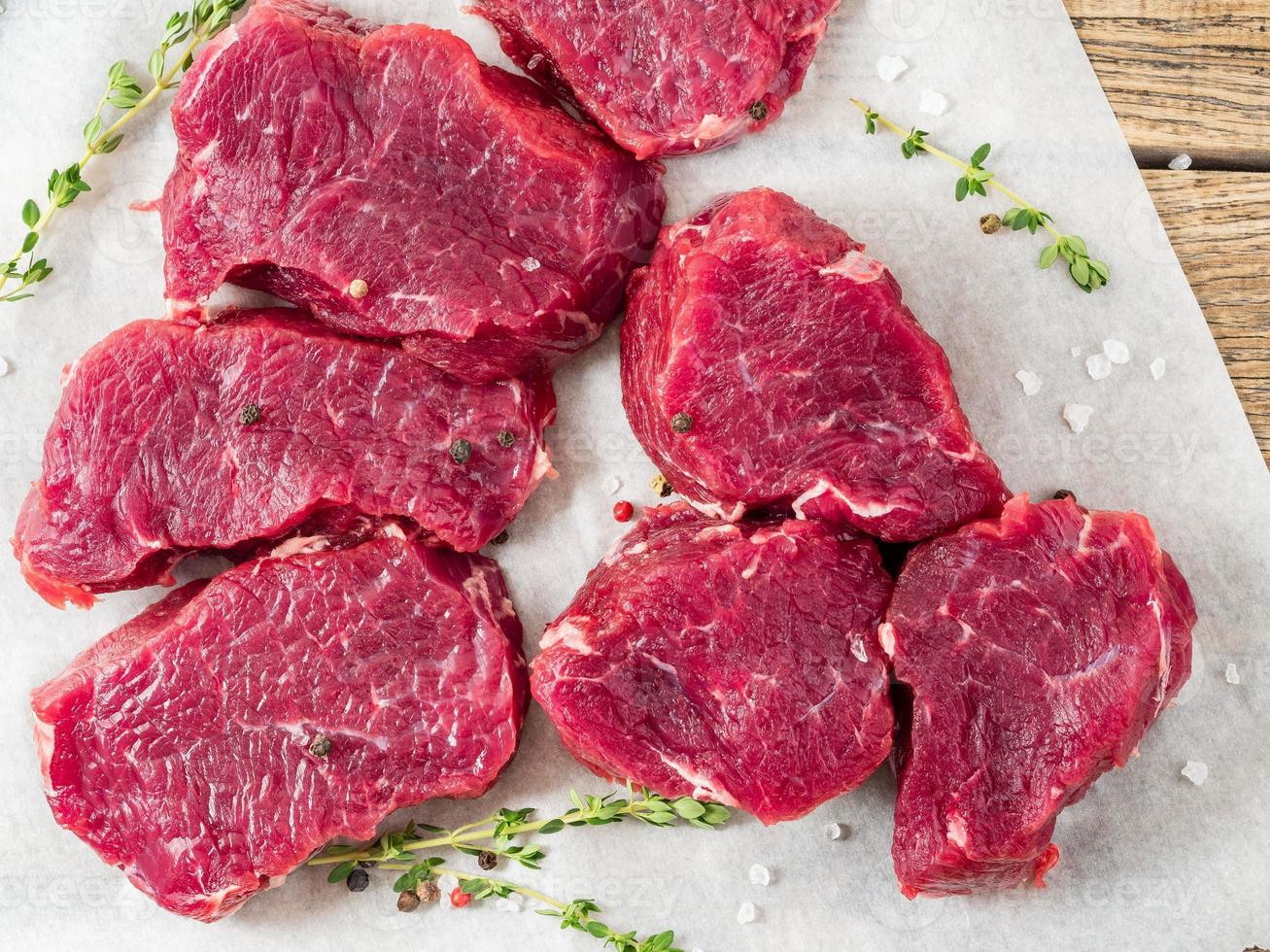 Stücke rohes Fleisch. rohes Rindfleisch mit Gewürzen und Thymian auf weißem Pergamentpapier auf rauem rustikalem Hintergrund, Draufsicht, Nahaufnahme. foto