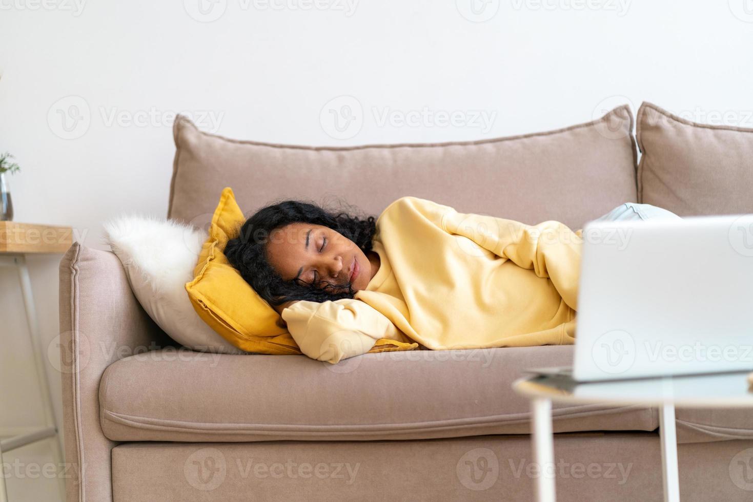 Gelangweilte afroamerikanische Frau, die auf der Couch im Wohnzimmer schläft, während der Film auf dem Laptop abgespielt wird foto