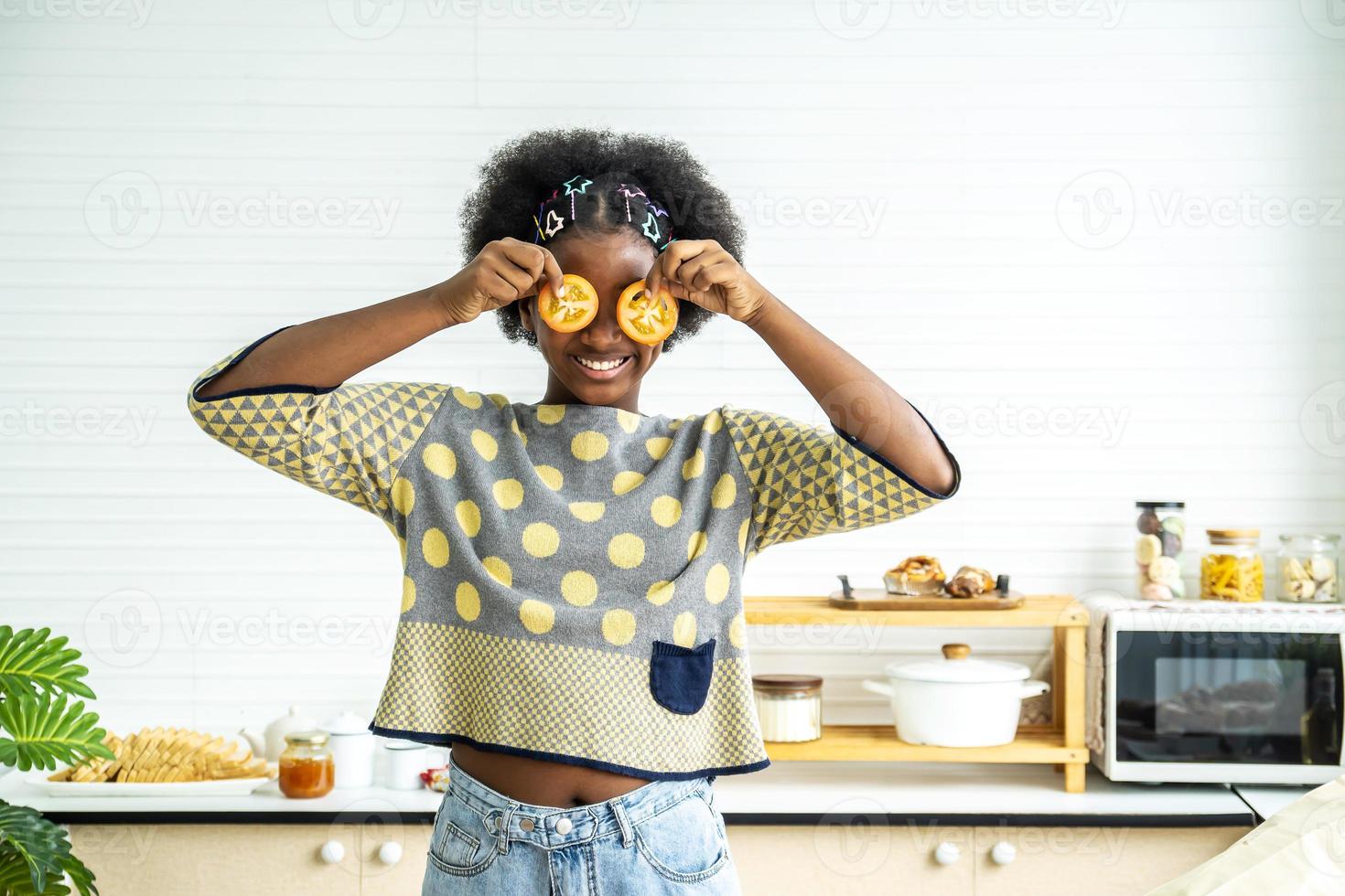 afrikanisch-amerikanisches glückliches mädchen, das sich in der küche mit lebensmittelgemüse vergnügt, hält tomaten vor seinen augen wie in einer brille und bringt dem kind gesunde und abwechslungsreiche vegetarische ernährung bei foto