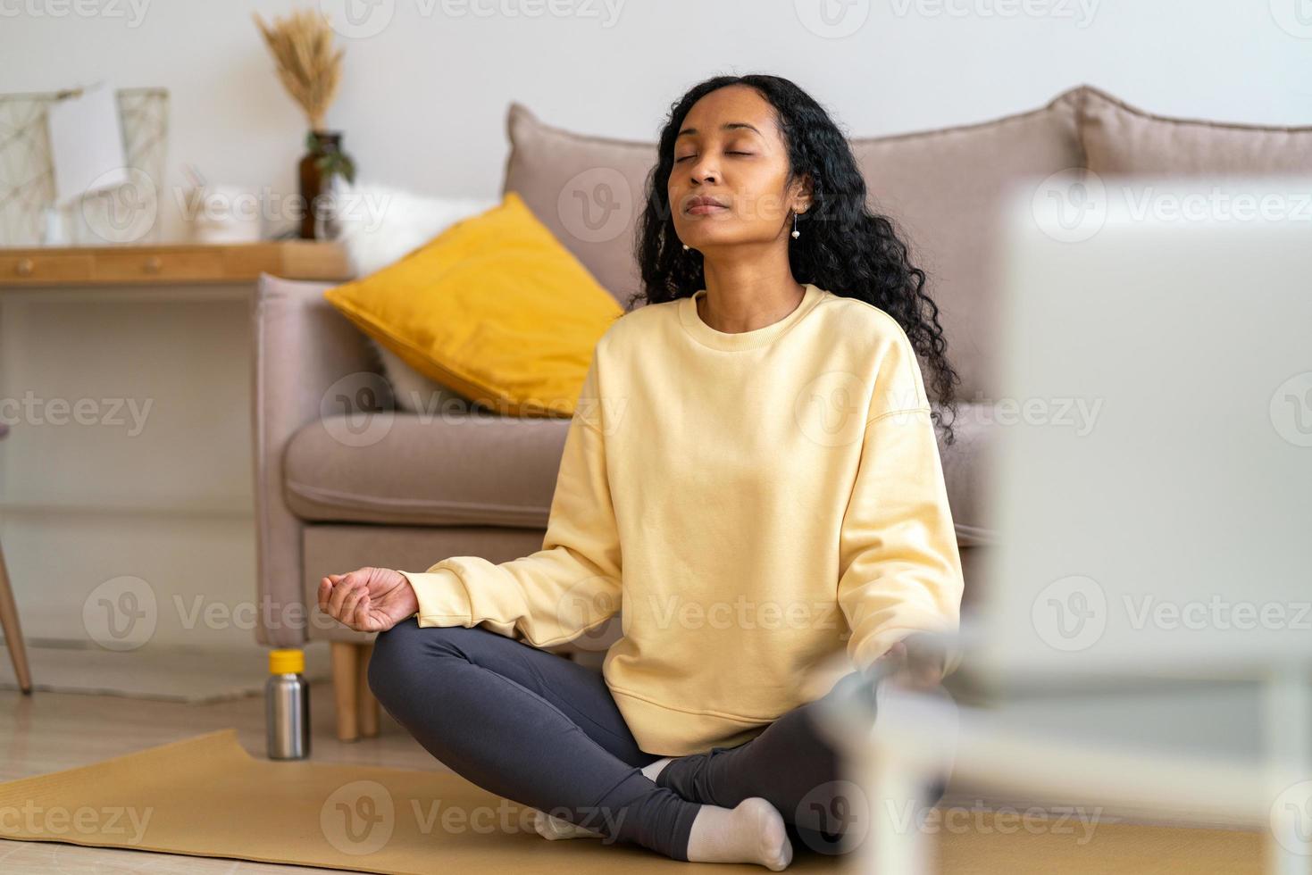 junge afroamerikanische frau, die in lotushaltung auf matte im wohnzimmer sitzt, während sie meditiert foto