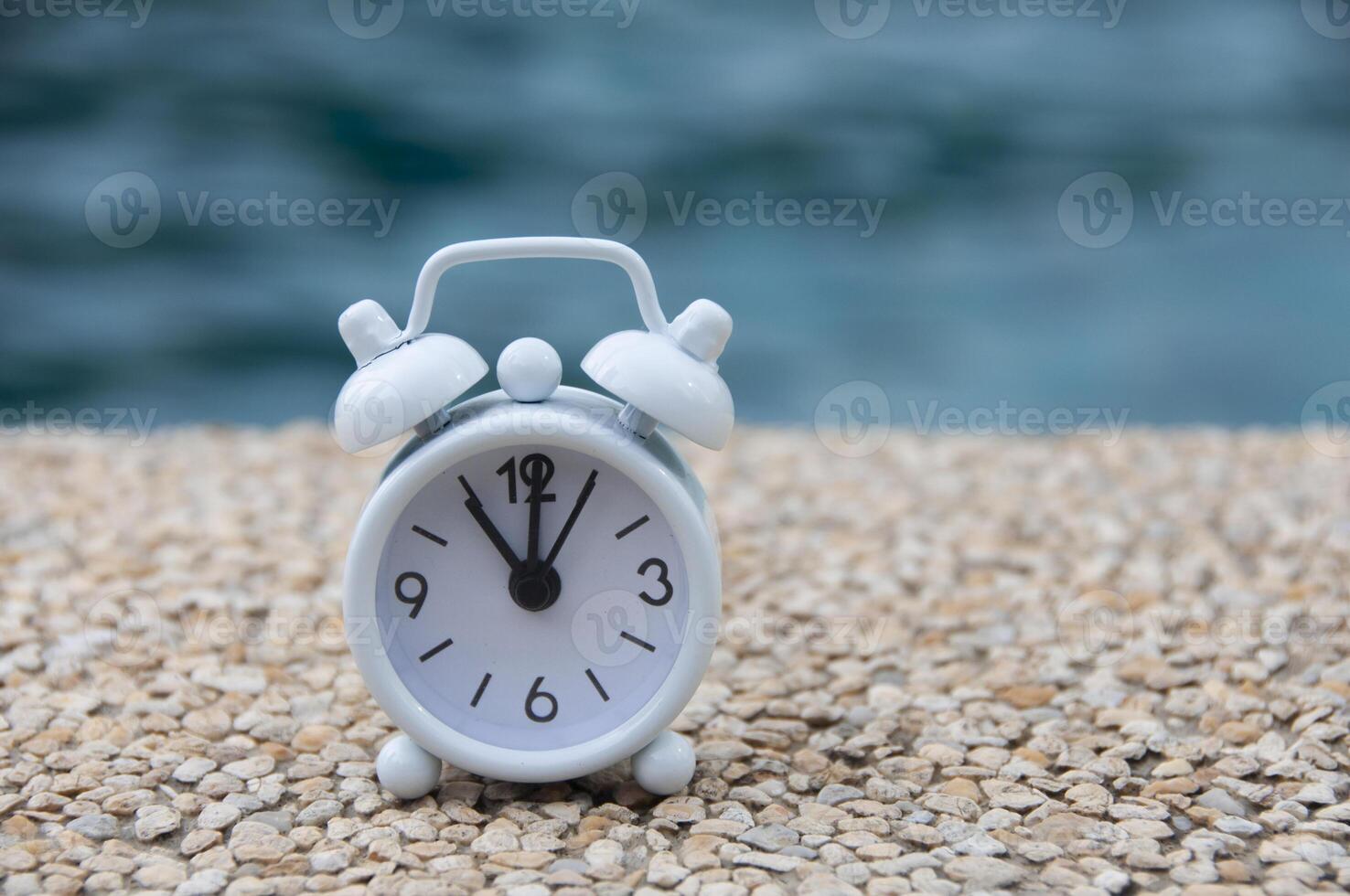 Weißer Wecker auf Marmorboden mit verschwommenem Swimmingpool-Hintergrund. die Uhr auf 11 Uhr gestellt. Terminkonzept. foto