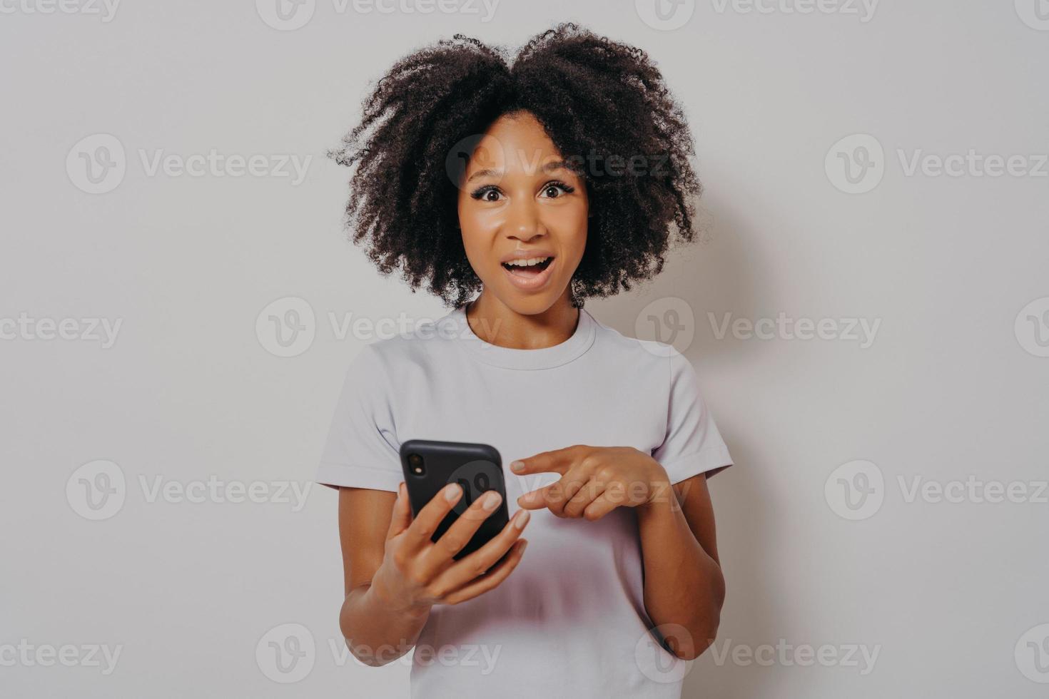 Aufgeregte, glückliche, dunkelhäutige Dame mit lockigem Haar, die ein Handy hält foto