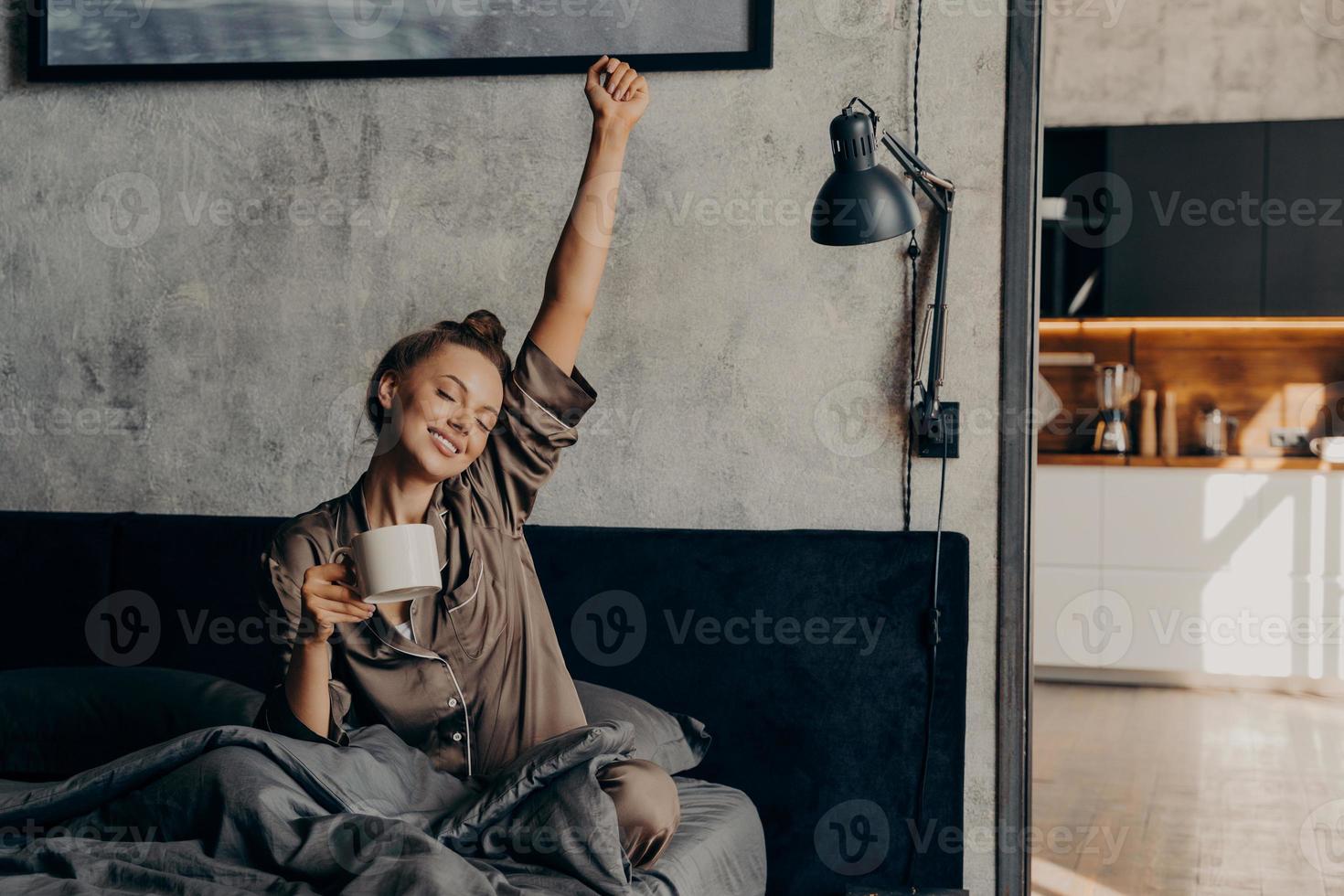 Schläfriges, entspanntes, hübsches Mädchen im Satinpyjama, das versucht, mit Kaffee aus dem Nachtschlaf aufzuwachen foto