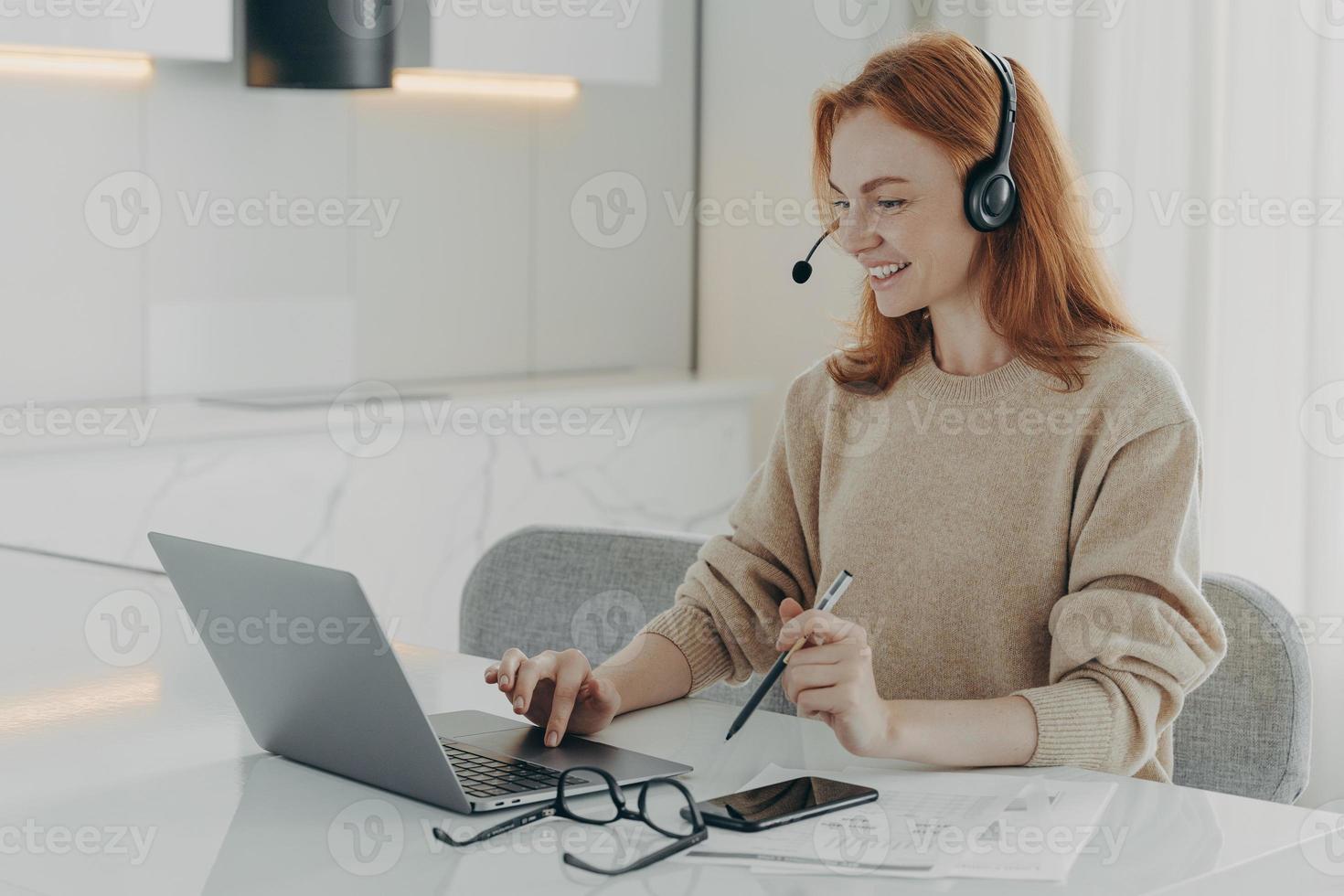 Glückliche rothaarige Frau sieht sich Online-Webinar an und verwendet ein Headset, das auf den Laptop-Bildschirm ausgerichtet ist foto