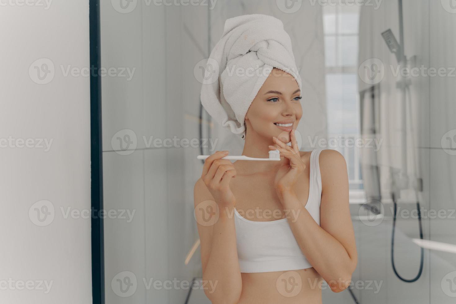 Mundhygienekonzept. Gesunde schöne Frau mit weißem Badetuch auf dem Kopf beim Zähneputzen während der Morgenroutine foto