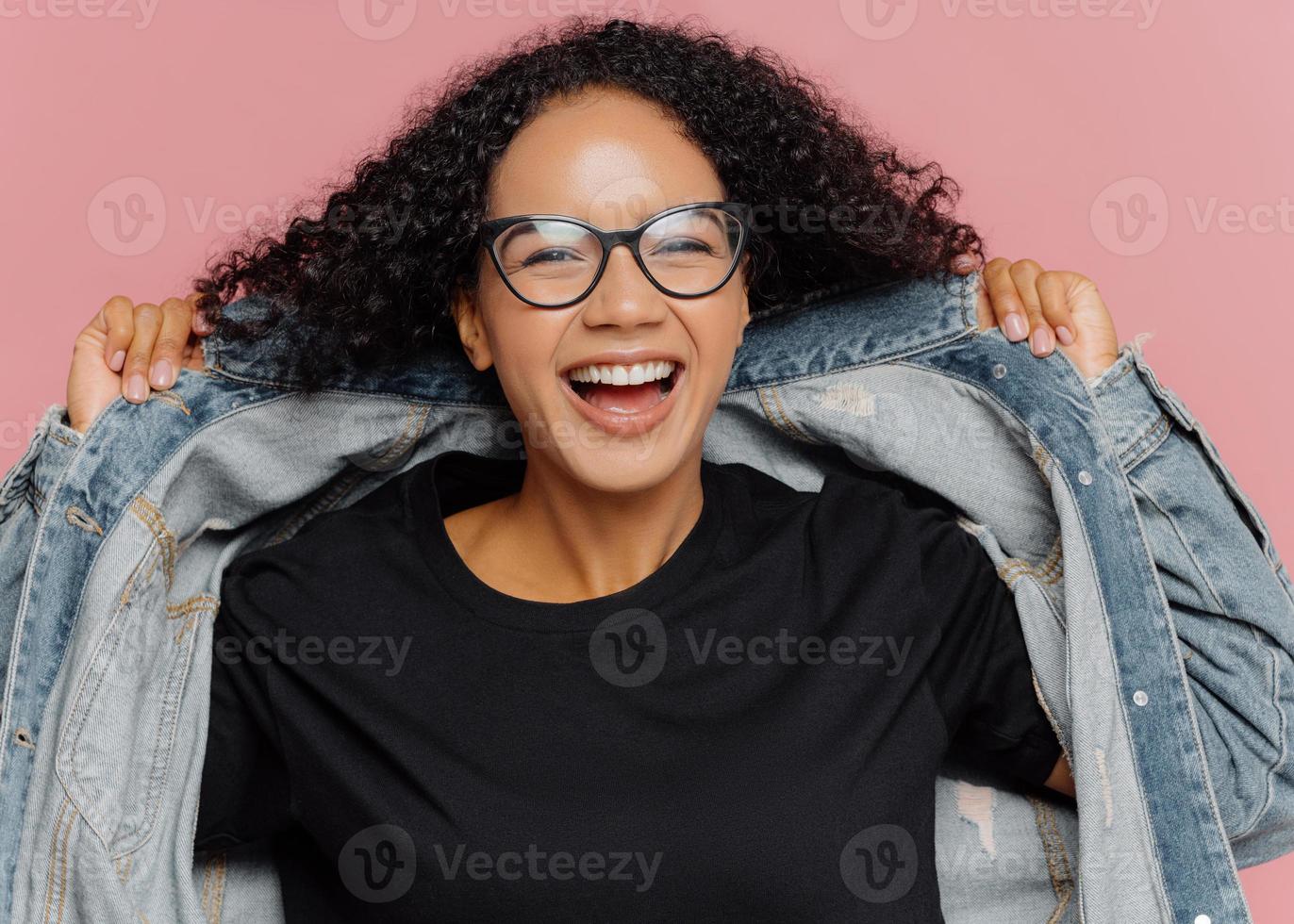 isolierte aufnahme einer lockigen, fröhlichen frau probiert eine neue jeansjacke an, trägt eine optische brille, lächelt breit, hat perfekte stimmung, posiert an einer rosa wand. positive Gefühle. Afro-Frau in stilvoller Kleidung foto