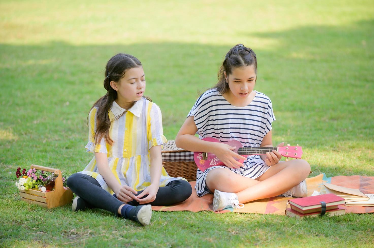 Zwei thailändisch-europäische Mädchen sitzen auf der Ukulele und singen, während sie im Park picknicken foto