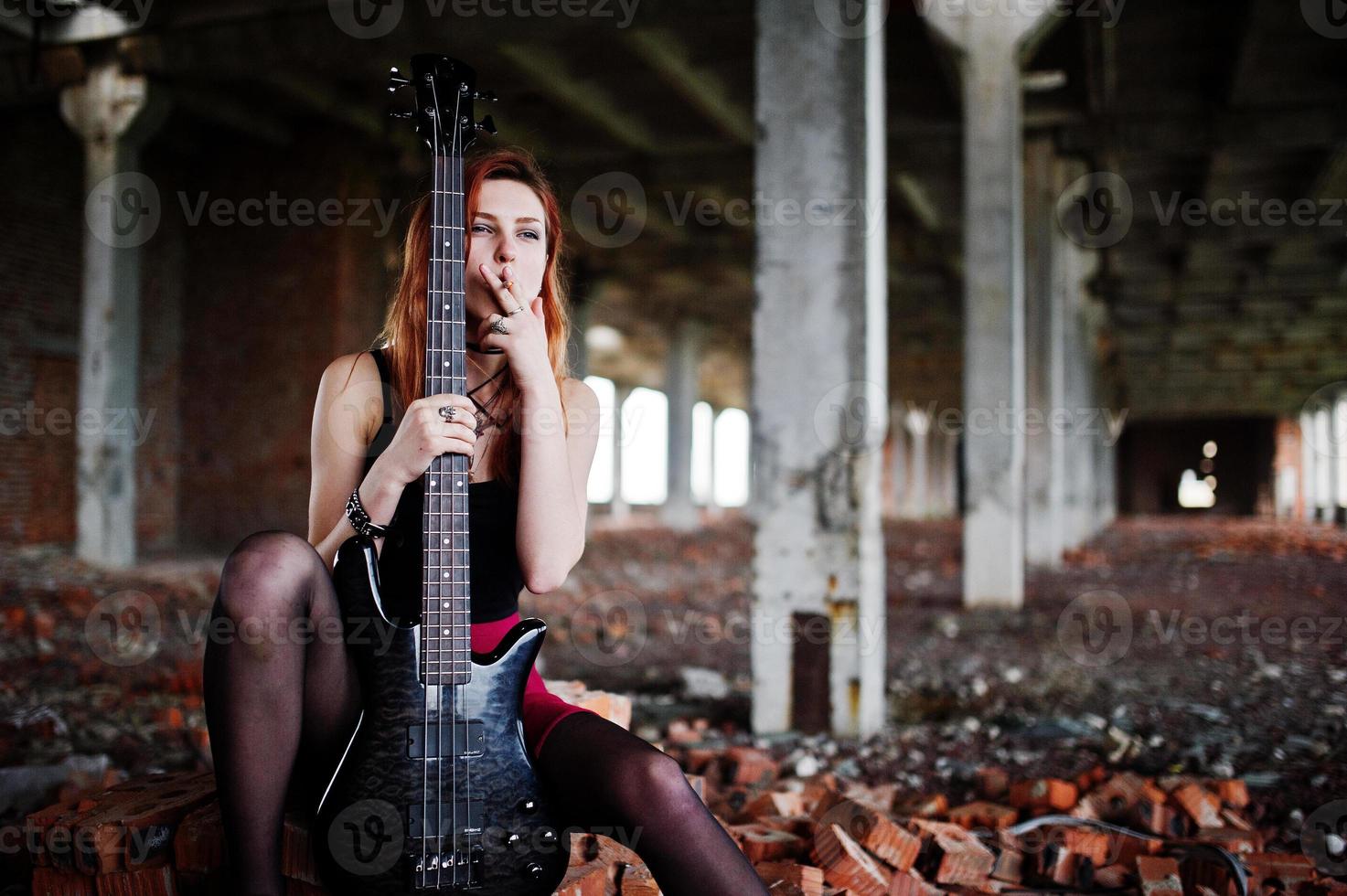 Rothaarige Punkmädchen tragen auf schwarzem und rotem Rock, mit Bassgitarre an einem verlassenen Ort. Porträt der gotischen Musikerin. Rauchende Zigarette. foto
