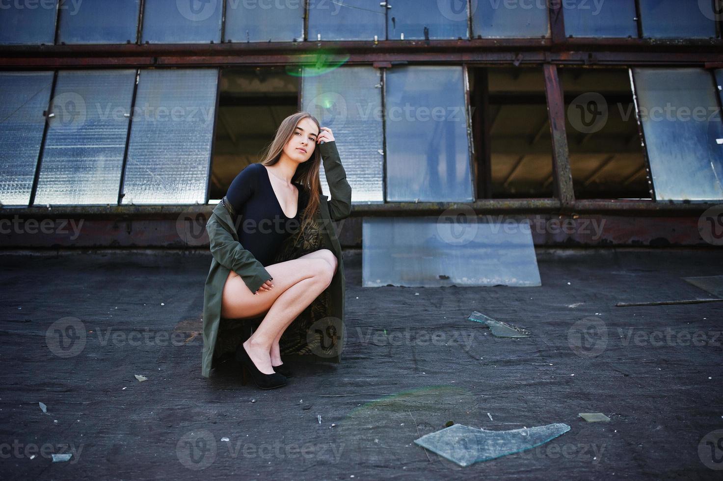 sexy Model-Mädchen mit langen Beinen im schwarzen Dessous-Outfit-Körper-Badeanzug-Kombikleid und Jacke posiert auf dem Dach eines verlassenen Industrieplatzes mit Fenstern. foto