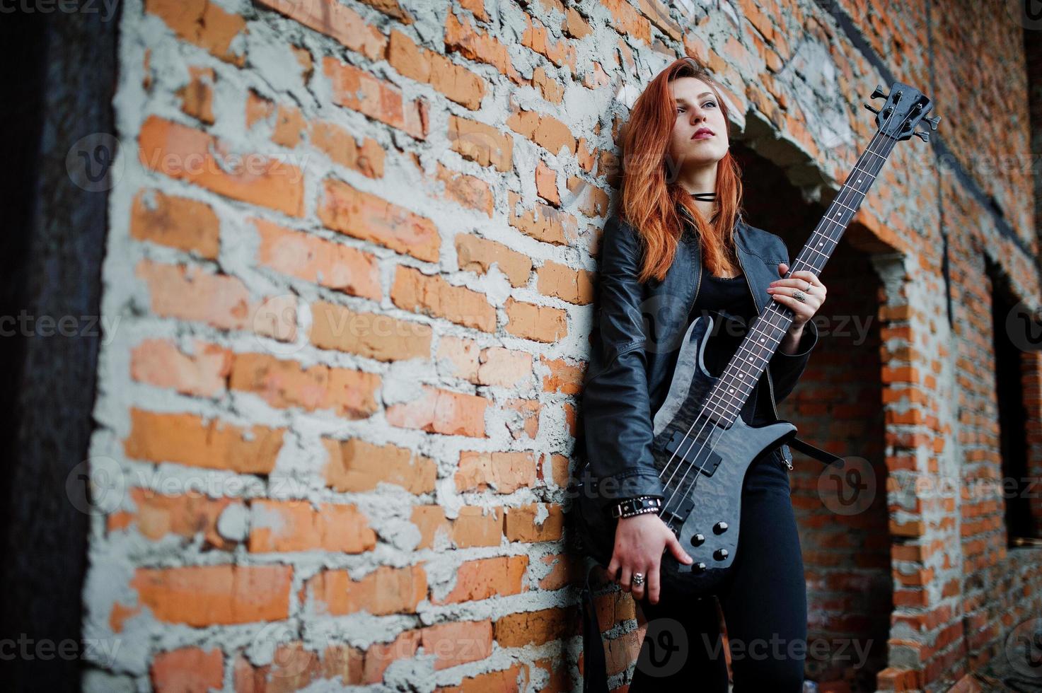 Rothaarige Punkmädchen tragen auf Schwarz mit Bassgitarre an einem verlassenen Ort. Porträt einer gotischen Musikerin. foto