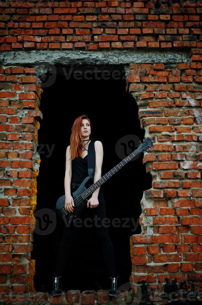 Rothaarige Punkmädchen tragen auf Schwarz mit Bassgitarre an einem verlassenen Ort. Porträt einer gotischen Musikerin. foto