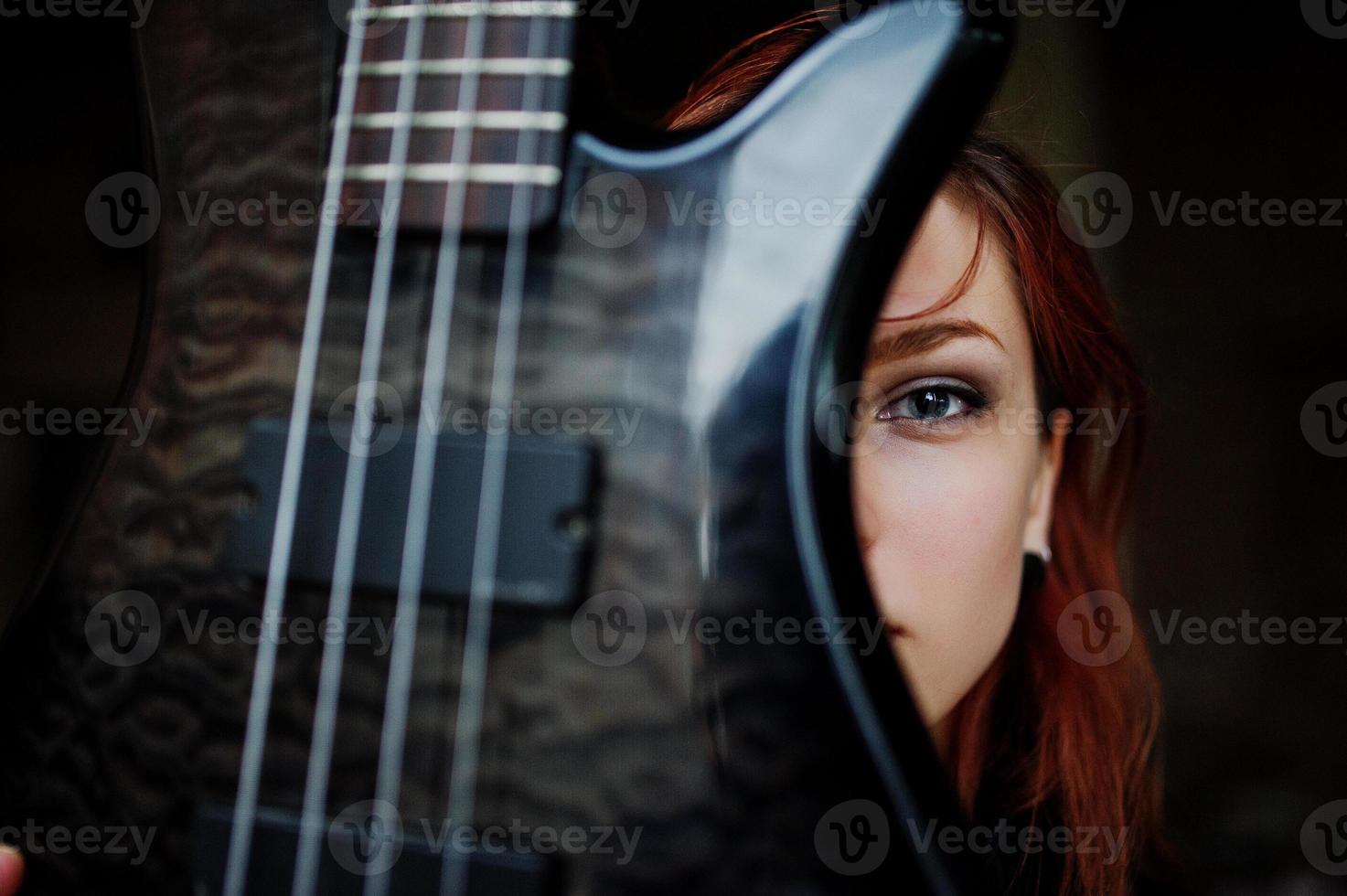 Rothaarige Punkmädchen tragen auf Schwarz mit Bassgitarre an einem verlassenen Ort. Porträt einer gotischen Musikerin. nahaufnahme gesicht der schwärze person mit gitarrenriff. foto